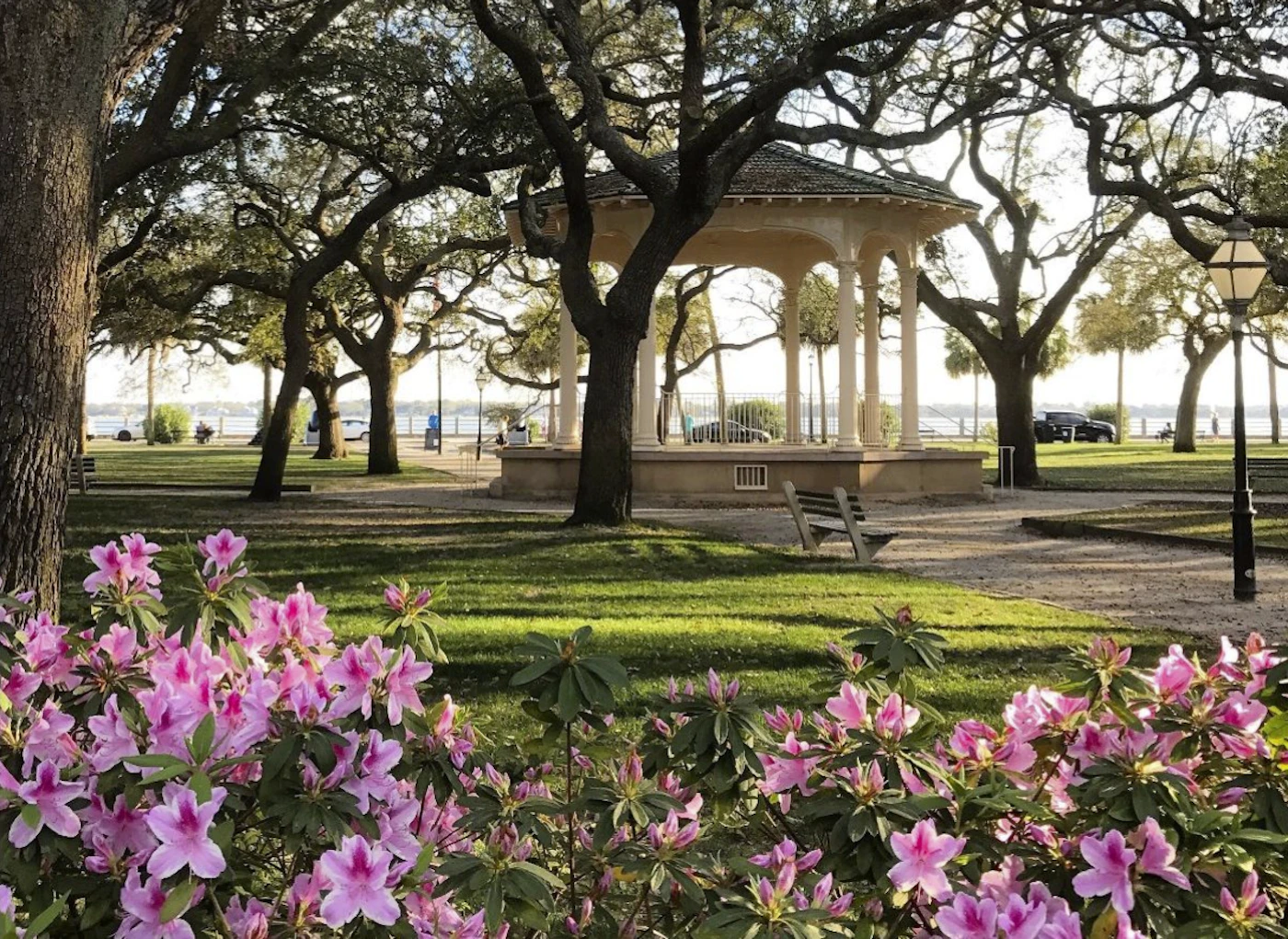 (Photo) A park scene featuring a gazebo, blooming azaleas, and large trees, with a body of water visible in the background. landscape | outdoor | gazebo, flowering bushes, trees, body of water Note: The image is a real-world photograph of a landscape scene. It captures a moment in time and depicts a natural setting.