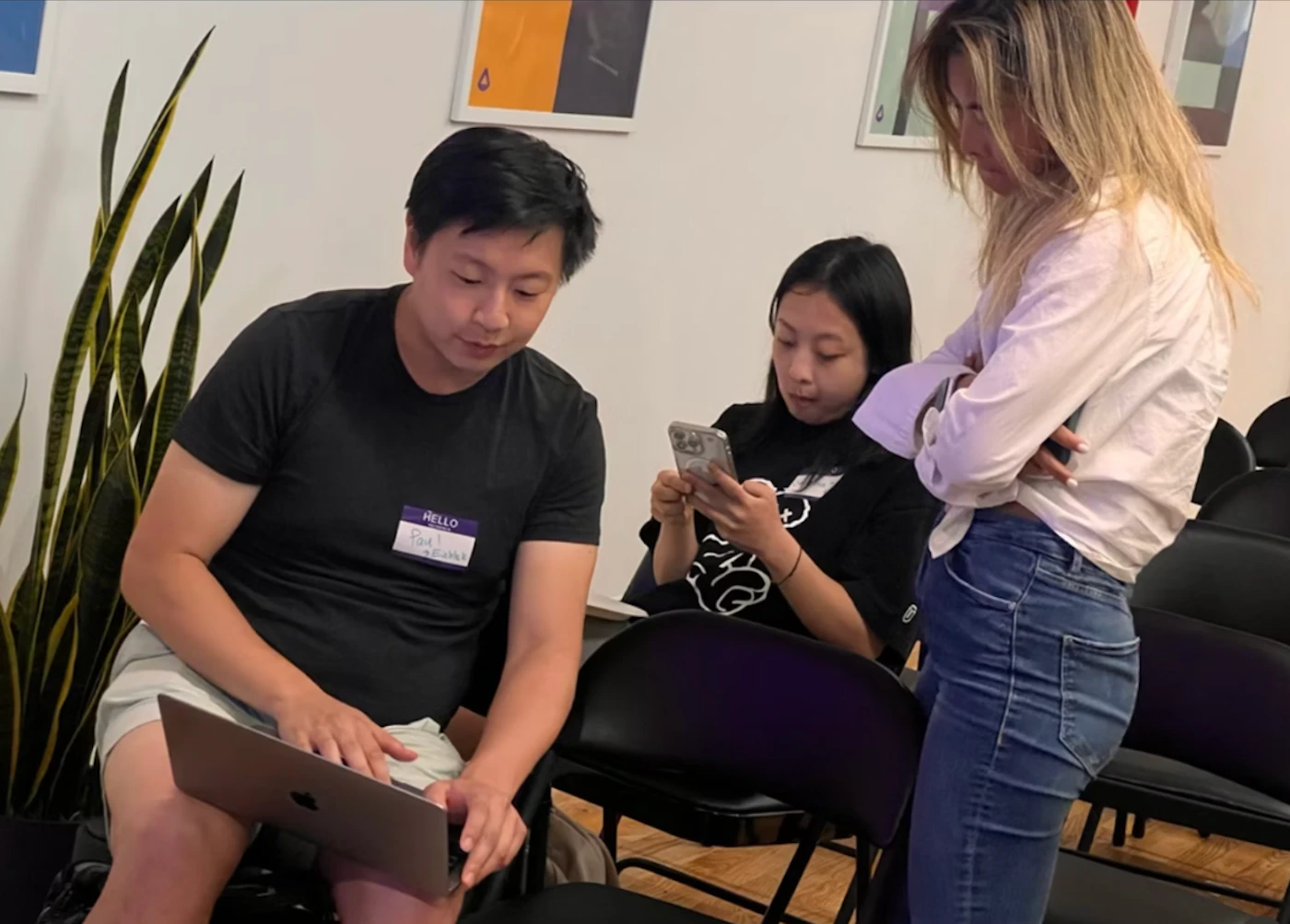 (Photo) Three people are gathered in a room, two are seated and one is standing.  One seated person is using a laptop, the other is using a phone, and the standing person has their arms crossed. Text: HELLO Paul [unreadable] people | indoor | three people, casual setting, laptop and phone use | candid Note: The image is a candid photograph of people in a room. It captures a real-life scene rather than a designed graphic or artwork.