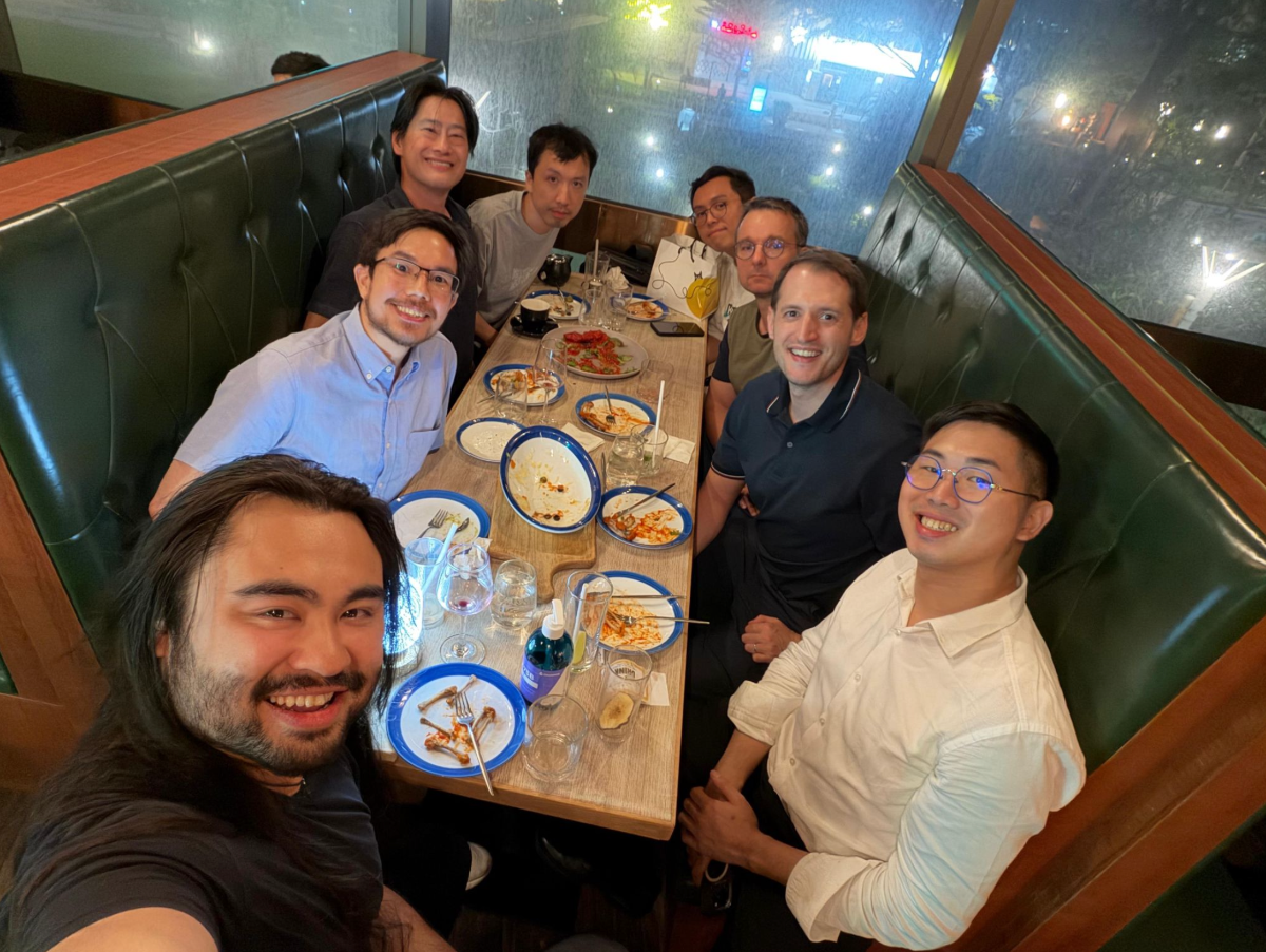 A group of eight men is gathered around a table in a dimly lit restaurant booth, smiling at the camera. The table is cluttered with dirty plates and glasses, suggesting they have just finished a meal.