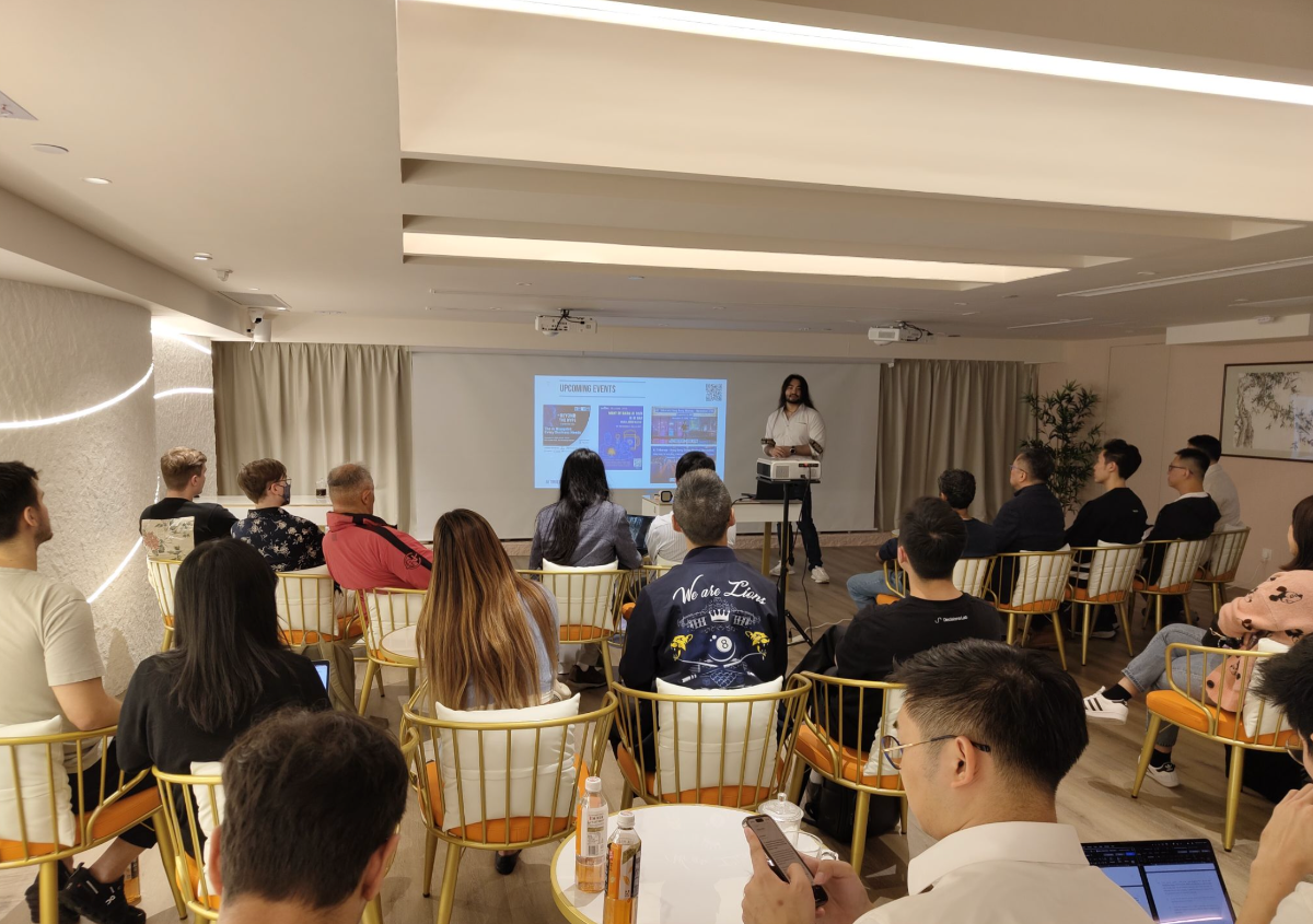 A photograph capturing an indoor event or presentation where an audience is seated in modern chairs, facing a projection screen that displays the title 'UPCOMING EVENTS.' A presenter stands to the right of the screen, addressing the group in a well-lit venue.. Text: UPCOMING EVENTS. We are Lions