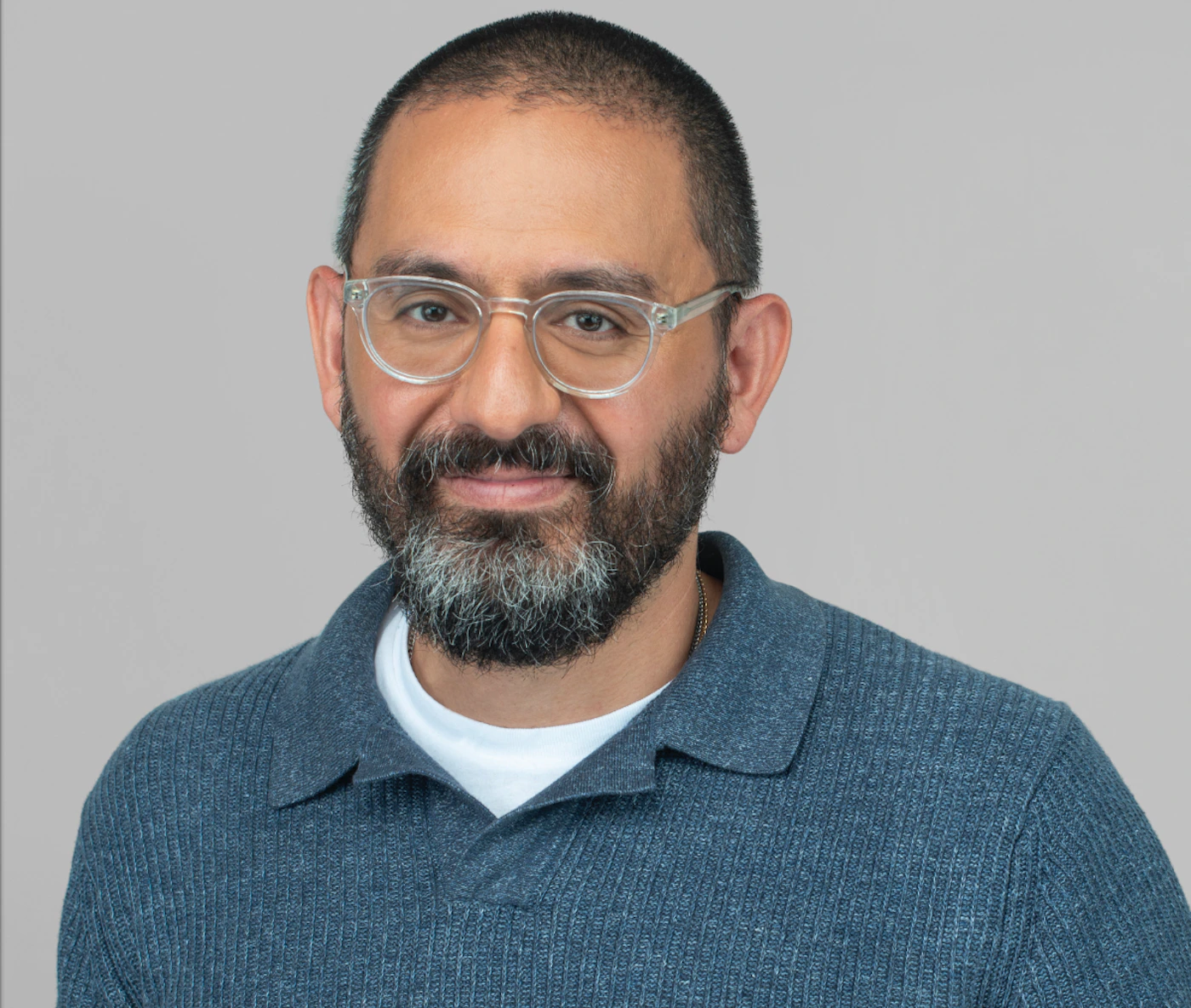 (Photo) A studio portrait of a man with a salt-and-pepper beard and clear-framed glasses, wearing a blue textured polo shirt against a plain gray background. person | indoor | man with a beard, clear-framed glasses, blue polo shirt | portrait | Colors: #B0B3B8, #4A6A8A, #2C2C2C Note: The image is a high-quality, realistic capture of a human subject, characteristic of professional portrait photography.