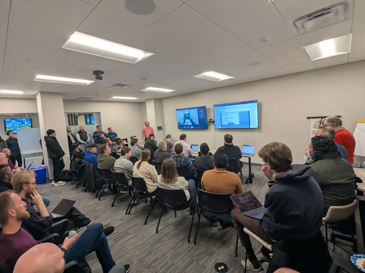 (Photo) A group of people are gathered in a conference room for a presentation, with two large screens displaying technical content and a Debian logo. people in a meeting or presentation | indoor office or conference room | multiple attendees, presentation screens, Debian logo, laptops | candid, wide-angle | Colors: #E0E0E0, #3C3C3C, #8B0000 Note: This is a real-life photograph capturing a group of people in an indoor setting during an event.