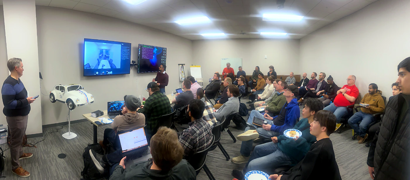 (Photo) A wide-angle photograph of a large group of people attending a tech-related presentation or demonstration in a conference room. Text: [unreadable] People attending a presentation | Indoor office or conference room | Two large wall-mounted displays, a model car on a stand, and many attendees with laptops. | Candid wide-angle shot | Colors: #f2f2f2, #2b2b2b, #004a99, #d11111 Note: The image is a real-life photograph capturing a candid scene of people in an indoor professional setting.