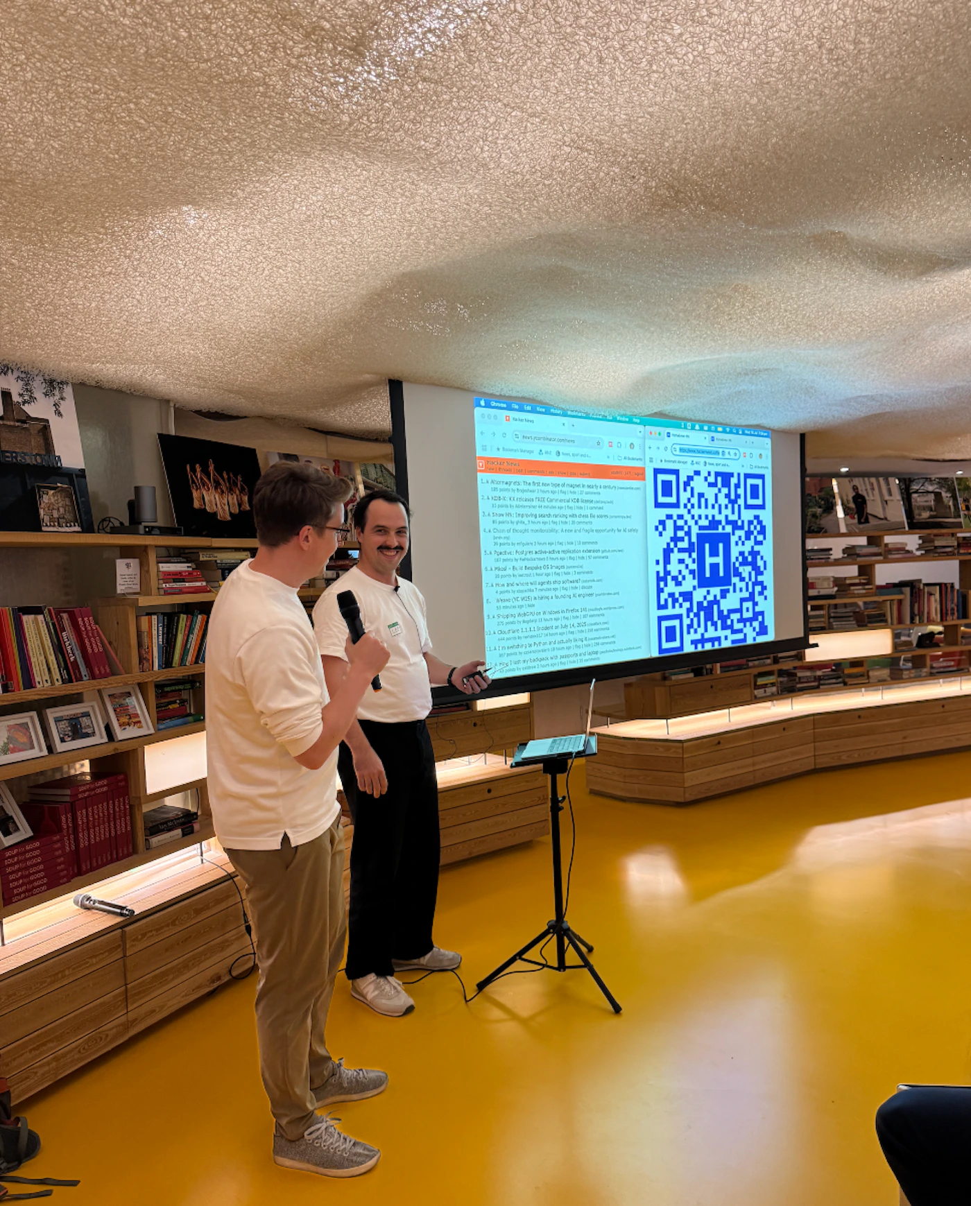 (Photo) The image captures two men giving a presentation or talk in a modern, brightly lit room with a distinctive textured ceiling and a vibrant yellow floor. A large screen behind them displays a list of points and a prominent QR code with an 'H' in its center. Text: HACKATHON 1.A Afterwards: The first ever type of [unreadable] in [unreadable] community 1.B [unreadable] and [unreadable] in [unreadable] of [unreadable] 2.A [unreadable] 90s, [unreadable] [unreadable] [unreadable] [unreadable] [unreadable] [unreadable] [unreadable] [unreadable] [unreadable] [unreadable] 3.A [unreadable] [unreadable] [unreadable] [unreadable] [unreadable] [unreadable] [unreadable] 4.A [unreadable] [unreadable] [unreadable] [unreadable] [unreadable] [unreadable] [unreadable] 5.A [unreadable] [unreadable] [unreadable] [unreadable] [unreadable] [unreadable] [unreadable] 6.A [unreadable] [unreadable] [unreadable] [unreadable] [unreadable] [unreadable] [unreadable] People giving a presentation | Indoor, likely an event space or communal area, characterized by bookshelves, a unique ceiling, and a bright yellow floor. | Two men (one holding a microphone), a large projection screen displaying text and a QR code with an 'H', extensive wooden bookshelves, and a textured, cream-colored ceiling. | Candid, event photography | Colors: #FDCB00, #F0EFE7, #9A7B5F, #007BFF, #000000 Note: This image is classified as a 'photo' because it is a direct capture of a real-world scene, featuring people, objects, and an environment in a naturalistic manner, characteristic of a photographic capture.