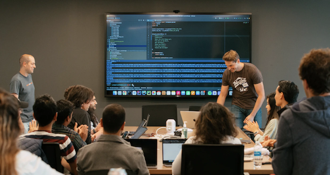 (Photo) A group of people are sitting around a table in a meeting room, looking at a large screen displaying code and applauding a presenter. people | indoor | meeting, presentation, code on screen, applause | candid Note: The image is a real-life captured moment of a group of people in a meeting room, making it a photograph. It depicts a scene rather than a designed graphic or piece of text.