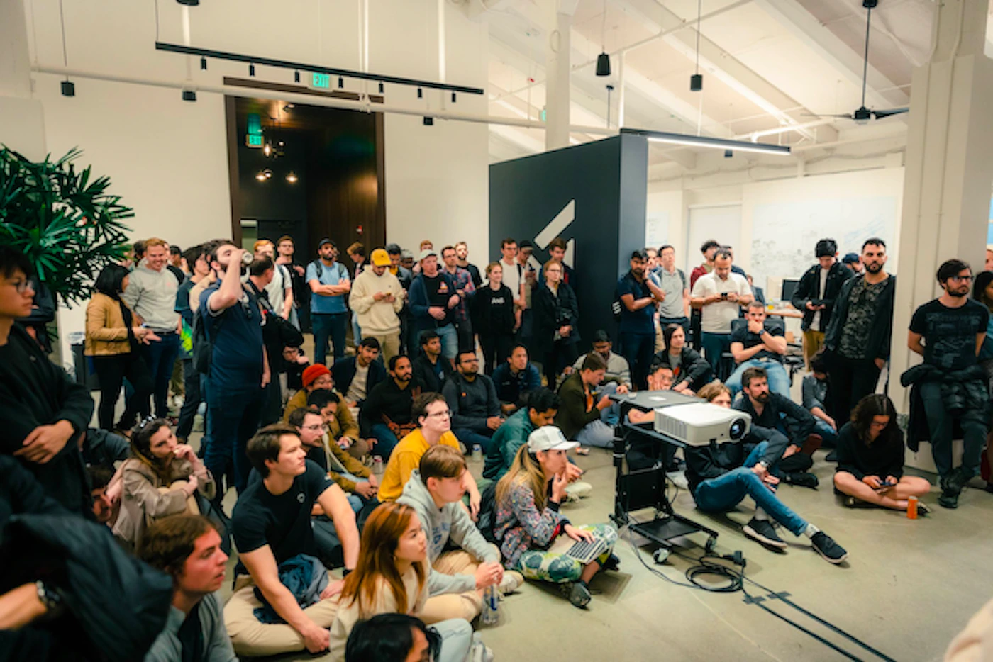 (Photo) A large group of people are gathered in a spacious room, many sitting on the floor, seemingly watching a presentation or event. A projector is set up in the center. people | indoor | large group, projector, presentation | candid Note: The image is a real-life captured moment of a gathering, indicative of a photograph. It's not a logo, illustration, banner, or handwriting.