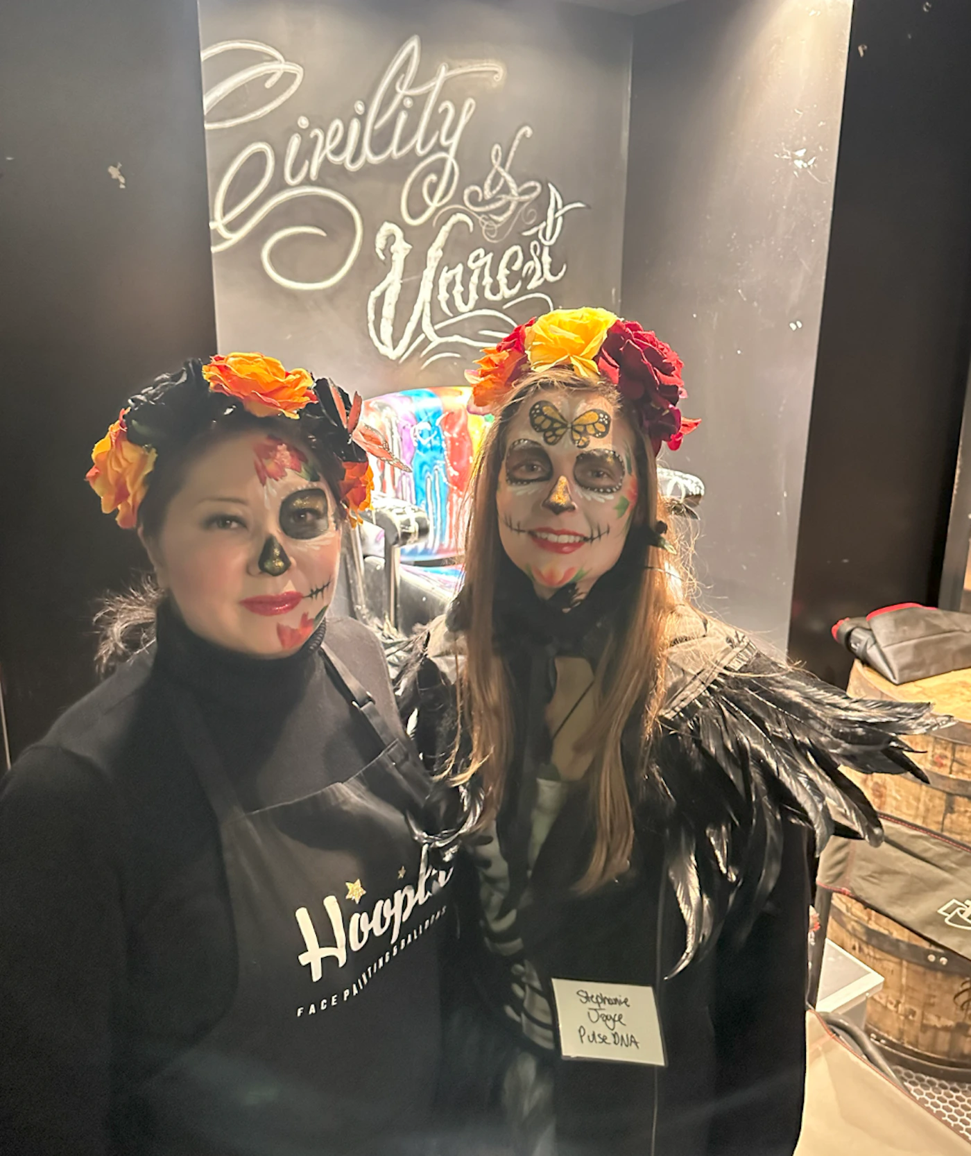 (Photo) Two people are wearing face paint and costumes, possibly for Halloween or Dia de los Muertos. They are standing in front of a wall with the words "Civility & Honest" written on it. Text: Civility & Honest Hoopla FACE PAINTING & BALLOONS Stephanie Dodge Pulse DNA person | indoor | costumes, face paint, two people | candid Note: The image is a real-life captured moment of two individuals, making it a photograph. It's not a staged promotional shot or an illustration.