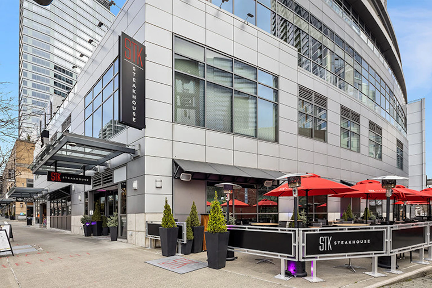 (Photo) A photo of the exterior of an STK Steakhouse restaurant, showing the building, outdoor seating area, and signage. Text: STK STEAKHOUSE STK STEAKHOUSE STK STEAKHOUSE building | outdoor | outdoor seating, signage | wide-angle | Colors: #d32f2f, #ffffff, #757575, #000000 Note: The image is a real-world photograph of a restaurant's exterior. It captures the scene as it is, rather than a stylized representation like a logo, illustration, or banner.