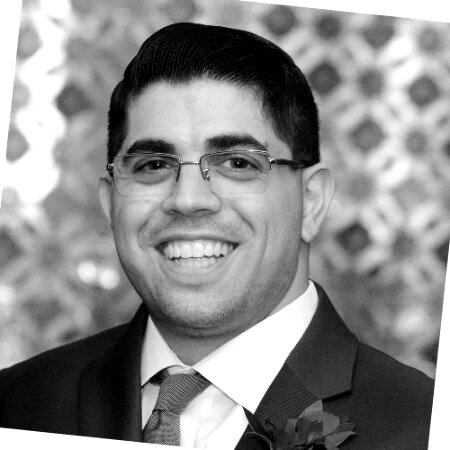 (Photo) A black and white portrait of a smiling man wearing glasses and a formal suit. person | indoor | smiling man, wearing glasses, formal suit and tie, patterned background | portrait, black and white | Colors: #000000, #FFFFFF, #7F7F7F Note: The image is a realistic capture of a person, characteristic of a professional headshot or portrait photograph.
