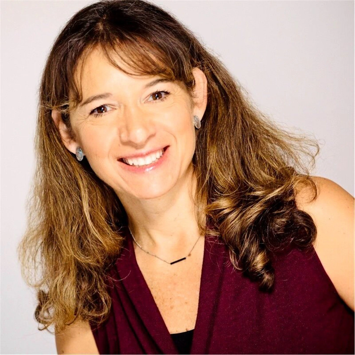 (Photo) A professional portrait of a smiling woman with long brown hair, wearing a maroon top and a simple necklace. person | indoor | smiling woman, studio lighting, professional attire | portrait | Colors: #FFFFFF, #4D1026, #8B613D Note: The image is a real-life photograph of a person, characteristic of a professional studio headshot.