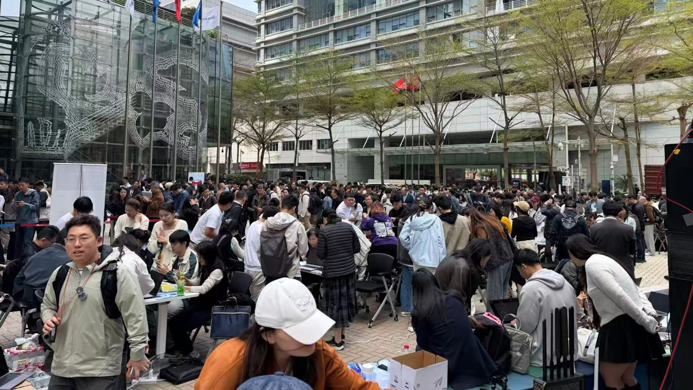 (Photo) A large, crowded outdoor event taking place in a modern urban plaza with people gathered around tables, many using laptops. large crowd, outdoor event, urban architecture | indoor | modern glass buildings, numerous people interacting at tables, laptops, urban trees | candid, wide-angle | Colors: #a9a9a9, #ffffff, #4a5d73, #8b4513 Note: The image is a real-life photograph capturing a candid scene of a large group of people in an outdoor setting.