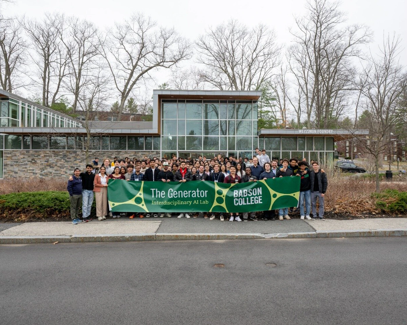 (Photo) A large group of people, likely students and faculty, stand outdoors on a sidewalk in front of a modern campus building, holding a long green banner. The banner promotes 'The Generator Interdisciplinary AI Lab' at 'Babson College.' Text: WEISSMAN FOUNDRY The Generator Interdisciplinary AI Lab BABSON COLLEGE Large group of people (students/faculty) and a modern institutional building. | Outdoor, on a college campus sidewalk, with bare trees in the background. | A large green banner with yellow text is held by the group; the building is identified as the Weissman Foundry. | Group portrait/candid | Colors: #A9A9A9, #4B5320, #FFFFFF Note: The image is a captured moment showing a large group of people posing outdoors in front of a building on a college campus, characteristic of a photograph.