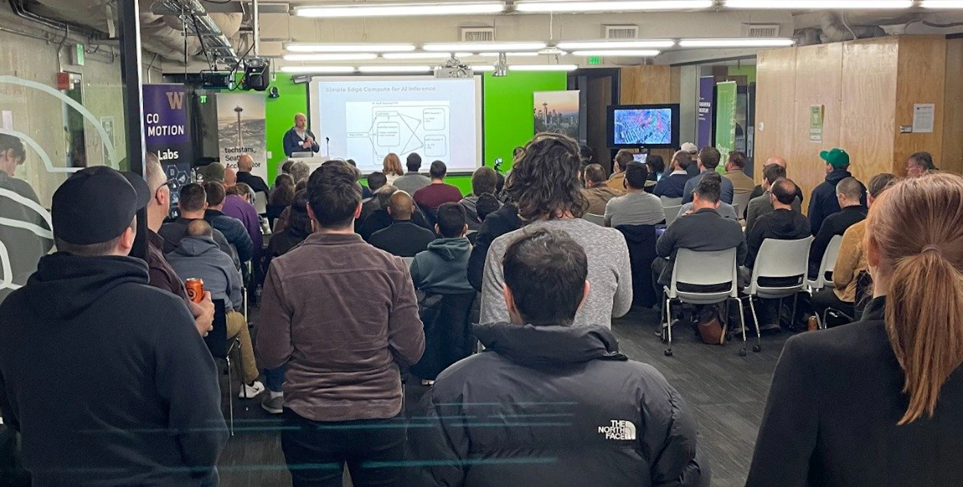 (Photo) A photo of an audience watching a presentation in a room. A presenter stands at a podium in front of a green screen, and the audience sits in chairs facing the presenter. Text: CO MOTION Labs people | indoor | presentation, audience, presenter | candid Note: The image is a real-life captured image of a presentation, depicting people and a setting. This makes it a photograph.