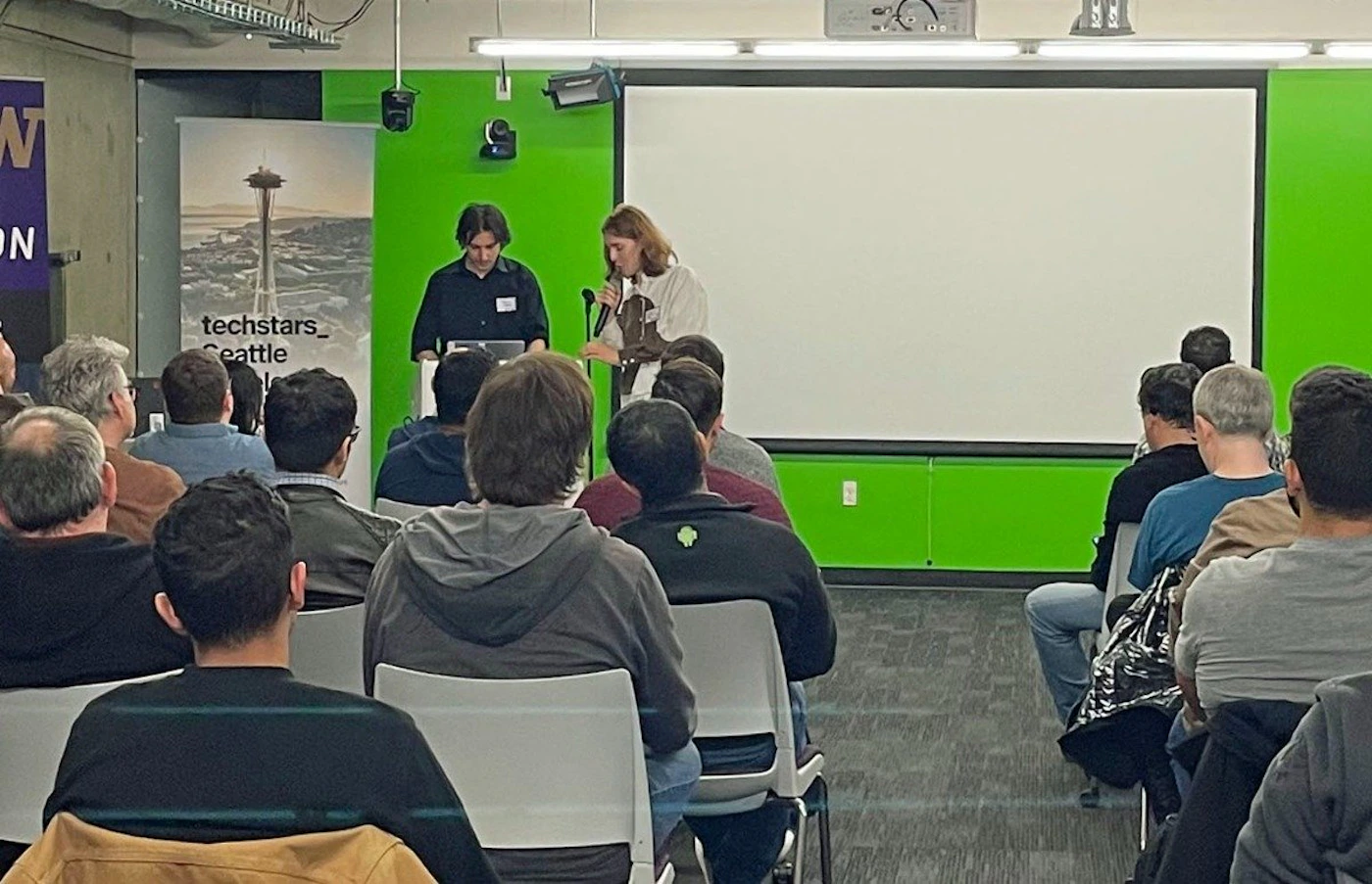 (Photo) A photo of a presentation taking place in a room with a green wall.  Two presenters stand at the front, while an audience sits in chairs facing them. Text: techstars_Seattle W ON people | indoor | presentation, audience, two presenters | candid Note: The image is a real-life captured image of a presentation, making it a photograph.  It depicts people in a presentation setting.