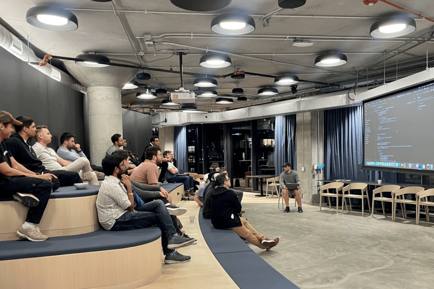 (Photo) A group of people are sitting in a modern office space, seemingly attending a presentation or training session.  One person is sitting on a chair with a laptop, possibly the presenter, while the others are seated on tiered, curved benches. people | indoor | group of people, presentation setting, modern office | candid Note: The image is a real-life captured scene of people in an office setting, indicative of a photograph. It's not a drawing, logo, or banner.