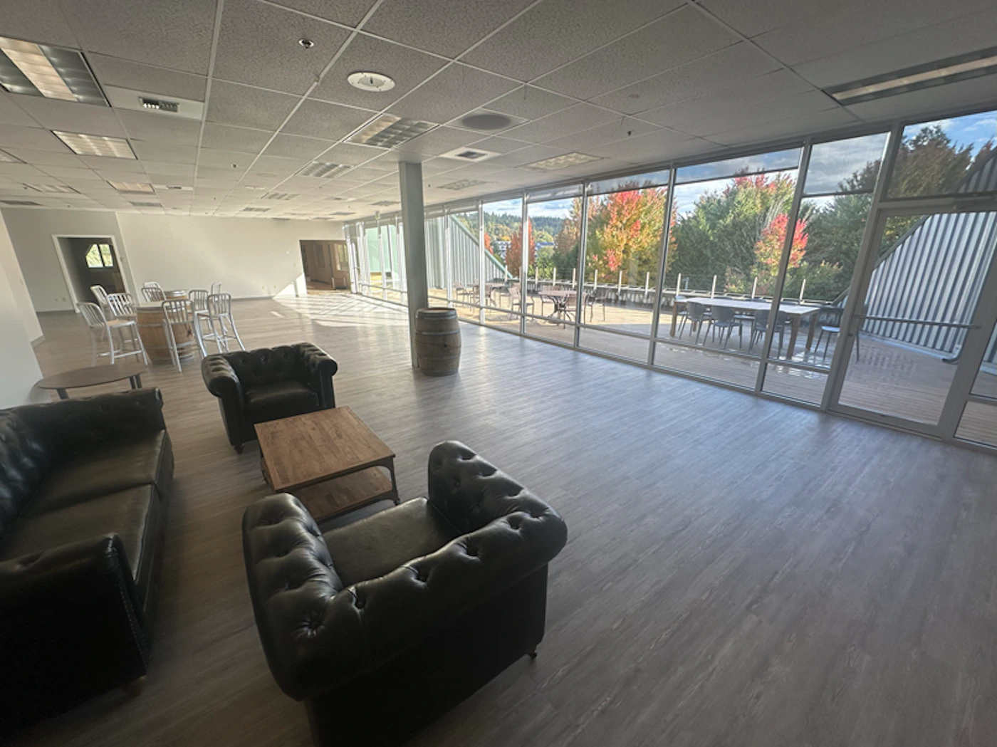 (Photo) A photograph showing the interior of a large, open commercial space or event room furnished with dark leather seating and wooden tables. A wall of floor-to-ceiling windows offers a view of an outdoor patio and trees displaying vibrant red and yellow autumn foliage. Interior commercial/event space and exterior landscape | Indoor (large room) looking out onto an outdoor patio, daytime | Large floor-to-ceiling windows, dark leather Chesterfield-style furniture, barrel tables, autumn foliage visible outside. | Interior architectural shot, wide-angle | Colors: #8c735c, #545454, #e0e0e0, #a04030 Note: The image is a realistic capture of an indoor commercial space looking out onto an outdoor patio and landscape, characteristic of a photograph.