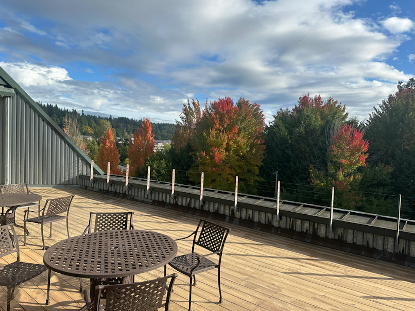 (Photo) A photo of an outdoor wooden deck furnished with dark metal tables and chairs, overlooking a dense wooded area displaying vibrant autumn foliage under a partially cloudy blue sky. outdoor patio/deck and autumn landscape | outdoor, likely a rooftop deck overlooking a forest or park | fall foliage (red, orange, green), dark metal patio furniture, wooden decking, corrugated metal siding on the adjacent building | scenic/landscape | Colors: #D2B48C, #8B4513, #FF4500, #4682B4 Note: The image is a realistic photographic capture of an outdoor scene, featuring patio furniture and a natural landscape.