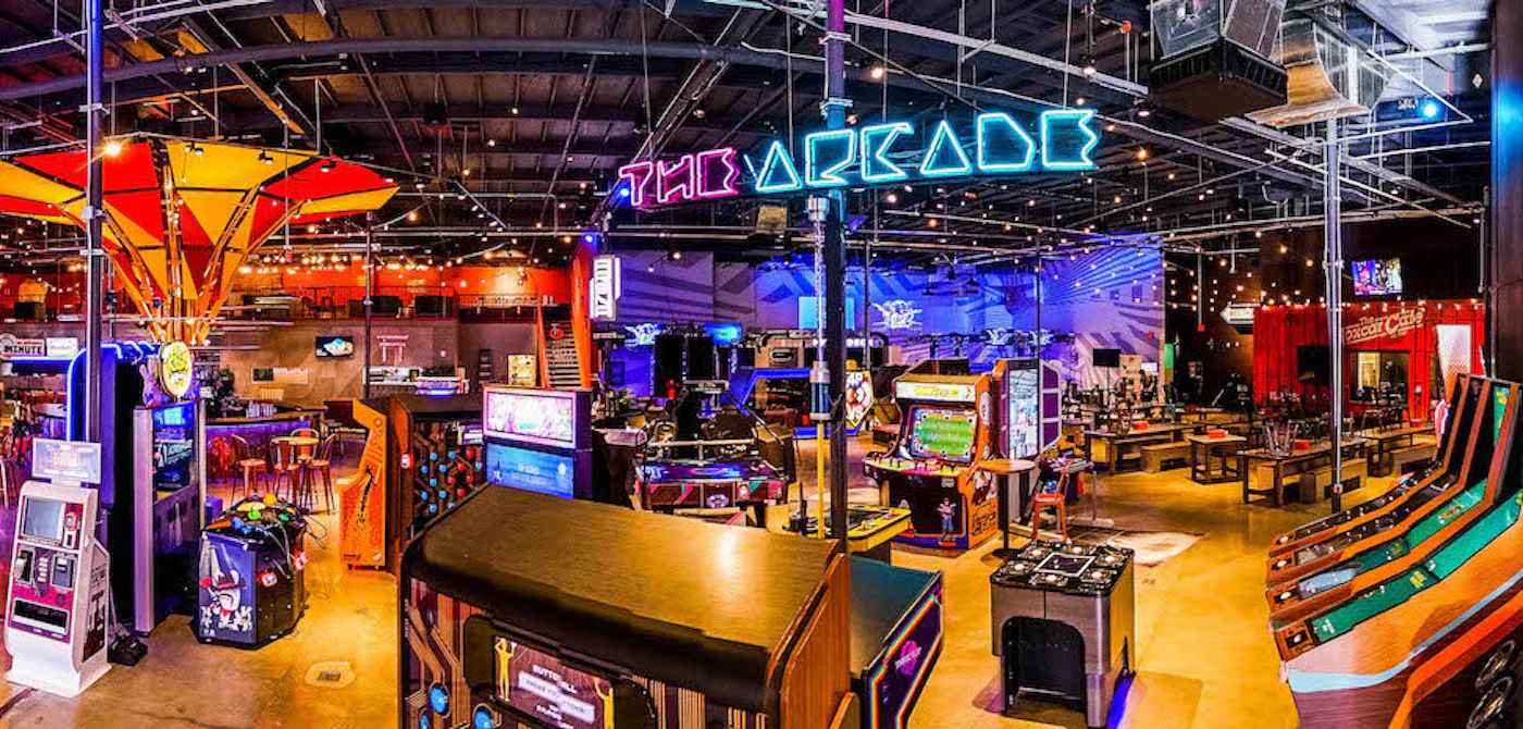 (Photo) A panoramic view of an indoor entertainment venue, likely an arcade, filled with various gaming machines and a bar area. Text: THE ARCADE indoor entertainment venue | indoor | arcade games, bar, neon sign | panoramic Note: The image is a real-world photograph of an arcade interior, not a digitally created image or a logo. It captures the scene as it is, showing the various elements and ambiance of the location.