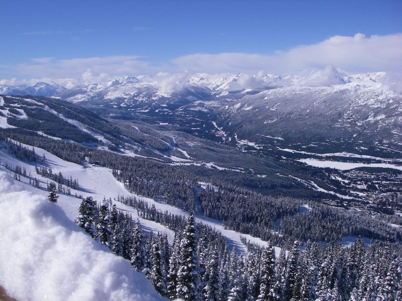 (Photo) A scenic vista of a snow-covered mountain range and valley, likely a ski resort, with evergreen trees lining the slopes. landscape | outdoor | snow-capped mountains, evergreen trees, ski slopes Note: The image is a real-world photograph capturing a natural landscape.