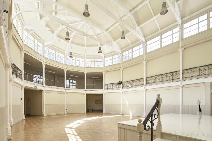 (Photo) A wide-angle photograph of a large, empty, circular hall with a high, vaulted ceiling and an upper balcony. The room features numerous windows along the upper walls, light-colored walls, a wooden floor, and a small stage or platform in the foreground. Text: 3D tagvenue architectural interior, event venue | indoor | high vaulted ceiling, large windows, circular layout, upper balcony, wooden floor, light-colored walls, stage/platform | architectural photography, well-lit, wide-angle | Colors: #E8E3D0, #FFFFFF, #B2926E, #8B8B8B Note: The image is a realistic depiction of an interior space, captured using a camera, indicating it is a photograph rather than an illustration, logo, or text-based format.