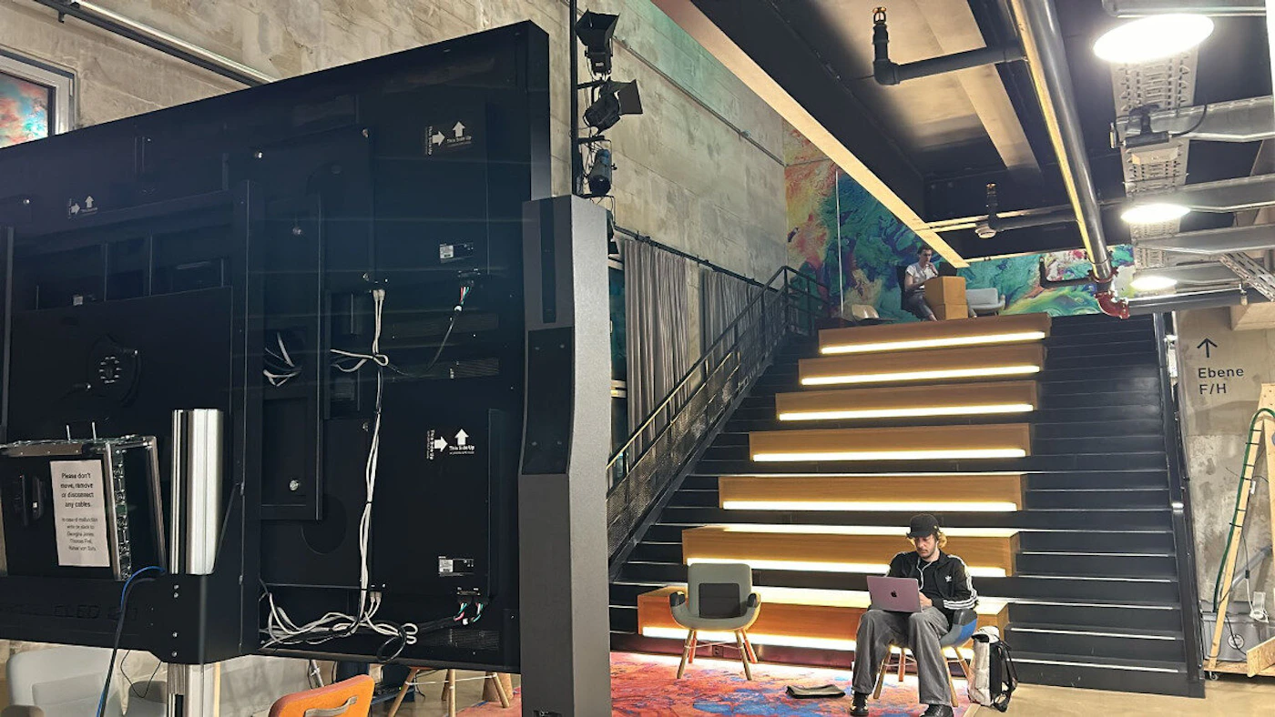 (Photo) A photo of the back of a large screen with visible wiring and a person sitting on a chair with a laptop near a staircase. Text: Please don't hang personal items on framed mirrors Though stylish, there is a risk of damage Indoor scene with person and objects | indoor | Large screen, wiring, staircase, person with laptop | candid Note: The image is a real-life captured scene of an indoor space, showing objects and a person in a specific environment. This makes it a photograph.