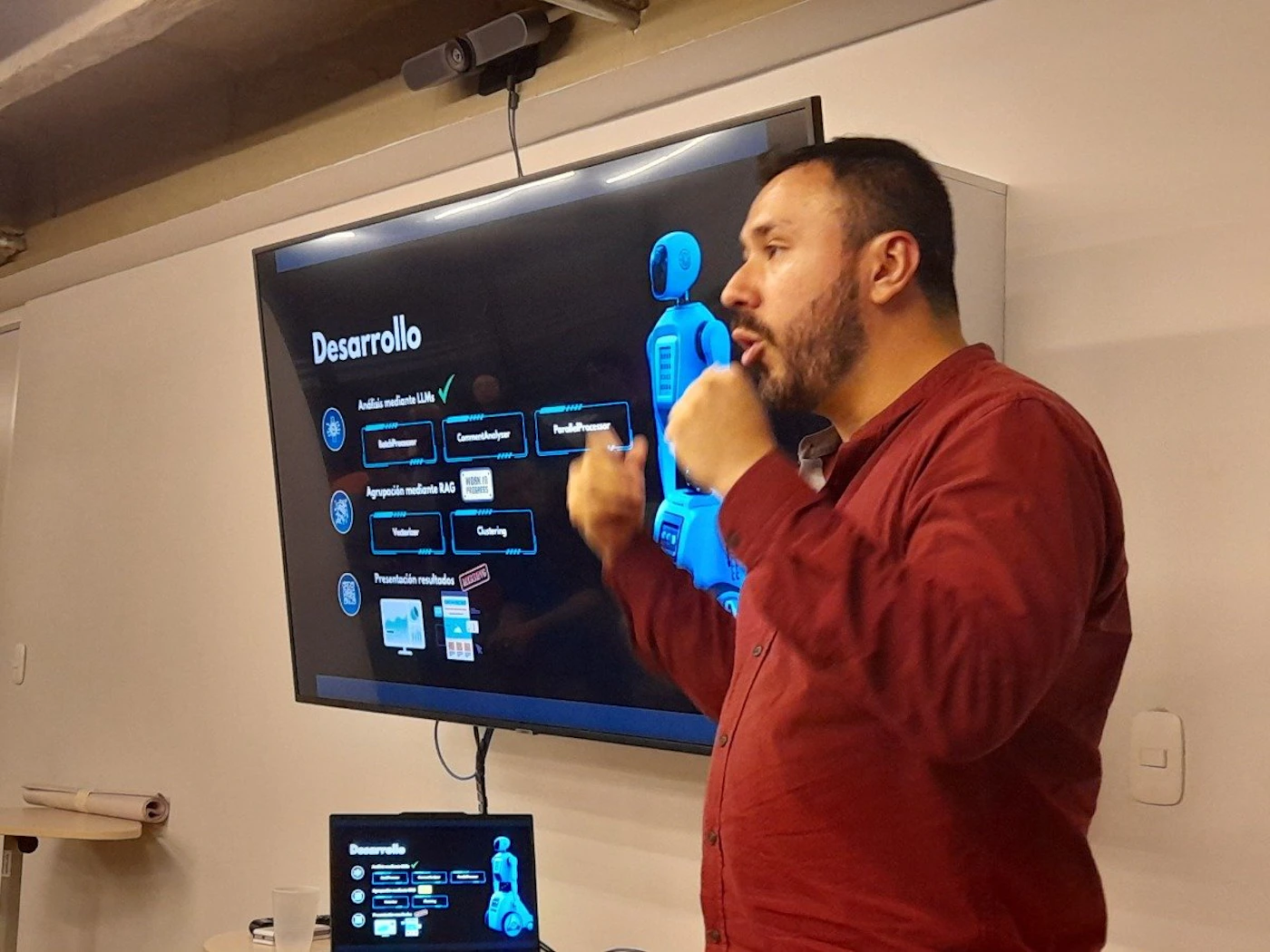 (Photo) A man gestures in front of a large screen displaying a presentation about software development. Text: Desarrollo Análisis mediante LLMs BuildProcessor CommandInterpreter PendfulProcessor Agregación mediante RAG Vectorizador Clustering Presentación resultados person | indoor | presentation screen, laptop | candid Note: The image is a real-life captured moment of a presentation, making it a photograph. It's not a staged setup for a logo, banner, or illustration.