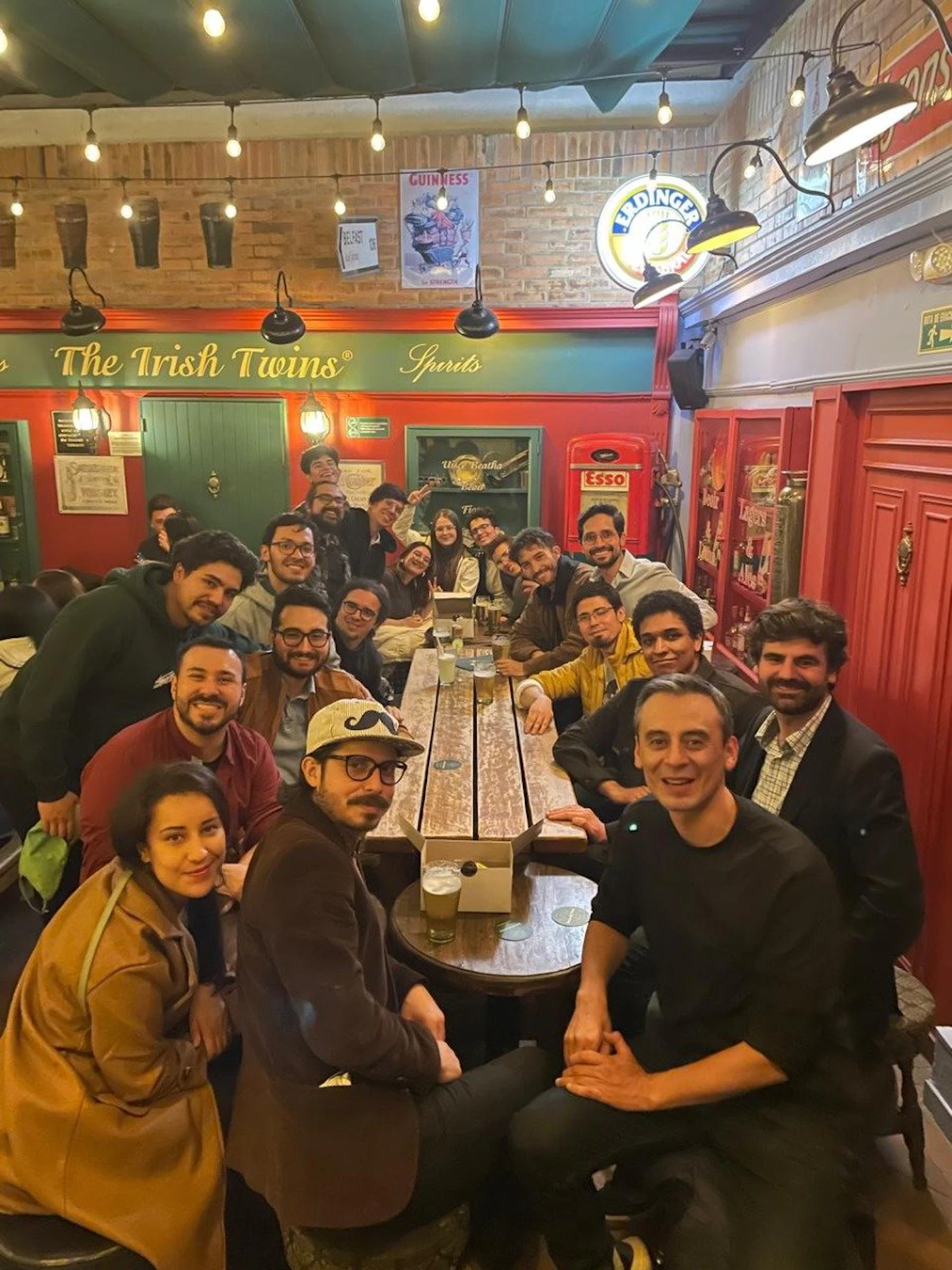 (Photo) A group of people are sitting together at a long table in an Irish pub, smiling for a photo. Text: The Irish Twins® Spirits GUINNESS ERDINGER Esso people | indoor | group photo, pub setting | candid Note: The image is a real-life captured moment of a group of people in a specific setting, indicating it's a photograph.