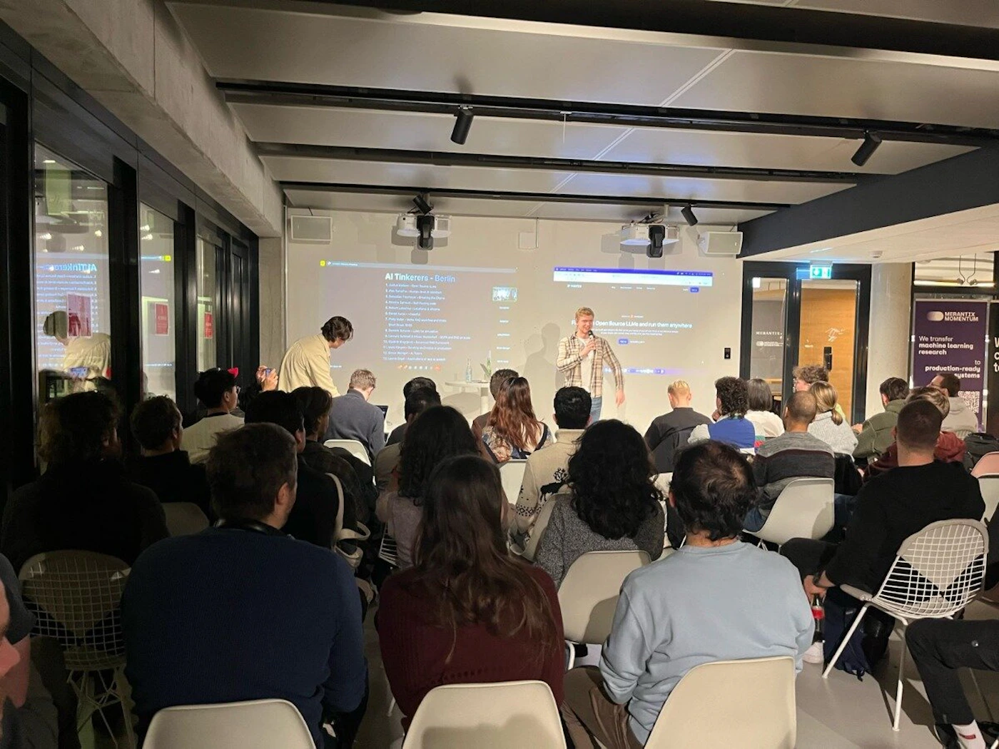 (Photo) A photo of a presentation taking place in a modern room.  An audience sits in chairs facing a speaker and a projected screen. Text: AI Tinkerer - Kotlin 1. What is an AI Tinkerer? 2. Why Kotlin? 3. How to get started? 4. Where to find help? 5. What are the benefits? Clear boundaries and how they are applied. MONTAJIA We transfer machine learning research to production-ready systems people | indoor | presentation, audience, projector screen | candid Note: The image is a real-life captured scene of a presentation, depicting people, a projector screen, and a room. This makes it best categorized as a photograph.