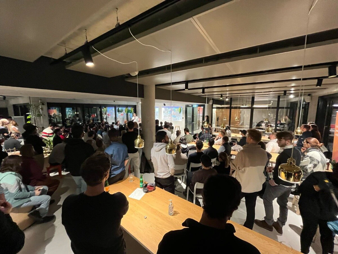 (Photo) A photo of a group of people gathered in a large room, seemingly attending a presentation or talk. people | indoor | presentation, audience, large room | candid Note: The image is a real-life captured scene of an event with people, indicating it's a photograph rather than a digitally created image or other types.
