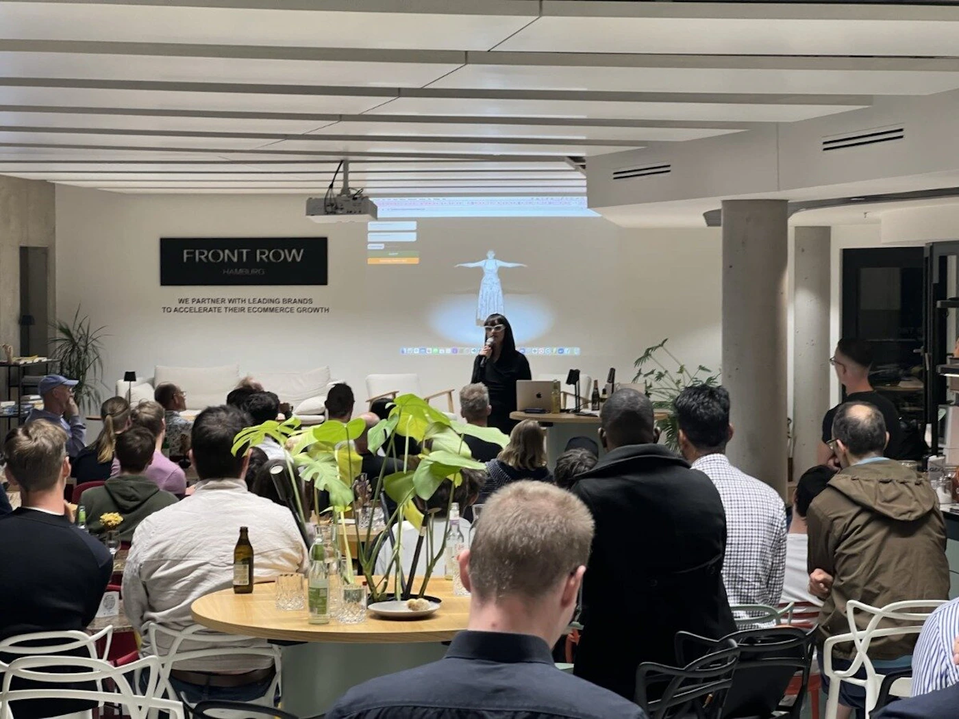 (Photo) A photo of a presentation taking place in a modern office space.  An audience is seated at tables, facing a speaker who is presenting with a projected image behind them. Text: FRONT ROW MAKES APS WE PARTNER WITH LEADING BRANDS TO ACCELERATE THEIR ECOMMERCE GROWTH people | indoor | presentation, audience, projector | candid Note: The image is a real-life captured scene of an event, making it a photograph. It depicts a presentation in progress.