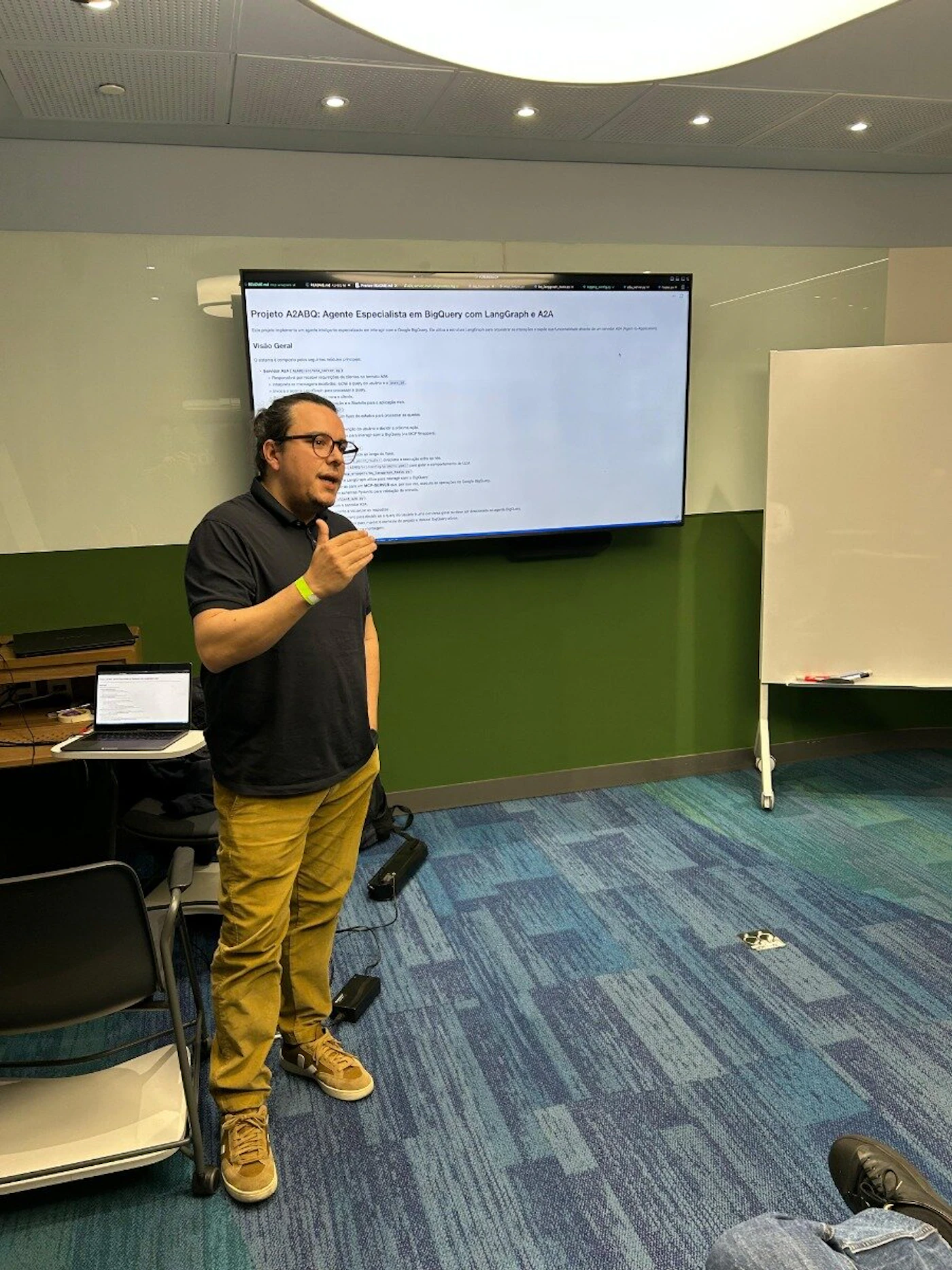 (Photo) A man wearing glasses, a dark polo shirt, and mustard-colored pants is presenting in a modern meeting room. He stands in front of a large screen displaying a presentation with text, and a white board is visible to his right. The room features a dark green wall and a patterned blue and teal carpet. Text: PROJETO A2ABQ: Agente Especialista em BigQuery com LangGraph e A2A Visão Geral O projeto é composto pela equipe [unreadable] e [unreadable] para o Google BigQuery. Ele utiliza uma LangGraph para [unreadable] e [unreadable] de um agente A2A. Preparação para o modelo [unreadable] para o BigQuery. Preparação para requisições de clientes na camada A2A. O modelo de BigQuery [unreadable] para o A2A. O BigQuery e LangGraph para [unreadable]. O BigQuery é a [unreadable] para o BigQuery. O LangGraph é um [unreadable] para o BigQuery. O A2A é um [unreadable] para o BigQuery. O A2A é um [unreadable] para o BigQuery. O A2A é um [unreadable] para o BigQuery. O A2A é um [unreadable] para o BigQuery. O A2A é um [unreadable] para o BigQuery. O A2A é um [unreadable] para o BigQuery. O A2A é um [unreadable] para o BigQuery. O A2A é um [unreadable] para o BigQuery. person presenting, indoor event | indoor, meeting room or classroom | man speaking with hand gestures, large screen displaying text, patterned carpet, whiteboard, laptop on a desk | candid, event photography | Colors: #38761D, #2B65EC, #B8860B, #212121, #F5F5F5 Note: The image captures a real-world scene featuring a person, a presentation screen, and a room environment. It is a direct photographic capture of an event.