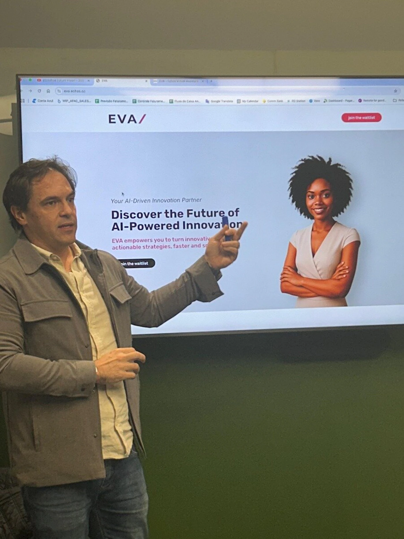 (Photo) A man is captured mid-presentation, standing in front of a large screen that displays a website or application interface. The screen features text related to AI-powered innovation, an image of a smiling woman, and 'join the waitlist' buttons. Text: EVA EVA (edited Virtual Assistant) Conta Azul WP_APAC_SALES... Previsão Faturame... Controle Faturame... Fluxo de Caixa An... Google Translate My Calendar Comm Bank Stripe Dashboard - Paga... Remote for good... EVA/ Your AI-Driven Innovation Partner Discover the Future of AI-Powered Innovation EVA empowers you to turn innovativ actionable strategies, faster and s[unreadable] join the waitlist join the waitlist A man presenting, a digital screen display, and a woman featured on the screen. | Indoor, likely an office or presentation room. | A large screen showing a modern website/application with text and a human image; a man pointing at the screen, suggesting a presentation or demonstration. | Candid, documentary. | Colors: #7A8072, #D7D0B7, #FFFFFF, #2E006A, #000000, #DBA483, #4C6C42, #E03A3C Note: The image is a real-world capture of a person in an indoor setting, interacting with a digital display. It documents a scene rather than being a standalone graphic or illustration.