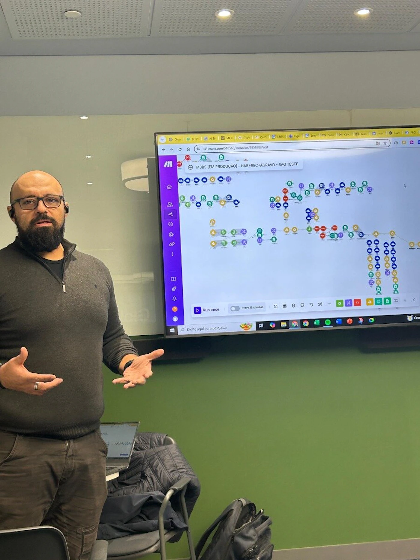 (Photo) A man with a beard and glasses is standing and gesturing in front of a large screen displaying a complex flow diagram or process map. The setting appears to be an indoor office or meeting room with a green wall. Text: s1.make.com/518566/scenarios/39580/edit M3BS EM PRODUÇÃO - HAB+REC+AGRAVO - RAG TESTE Run once Every 15 minutes Digite aqui para pesquisar person, presentation screen, meeting room | indoor | man gesturing, large digital display with a flowchart, laptop and backpack visible | candid, presentation | Colors: #4CAF50, #333333, #FFFFFF, #800080, #F0F0F0 Note: The image is a realistic capture of a scene, featuring a person and a presentation screen in a real-world environment, which is characteristic of a photograph.