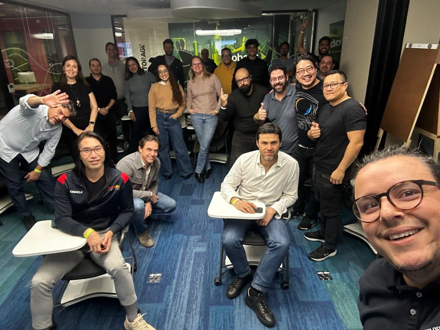 (Photo) A group of diverse individuals, mostly adults, are gathered in what appears to be an office or classroom setting, posing for a photo. Many are smiling and looking towards the camera, with some making gestures like thumbs up or waving. Text: STORAGE ORACLE oba group of people | indoor, likely an office or classroom | multiple people, some smiling and gesturing, desks and chairs visible, a banner and wall art in the background | group portrait, candid | Colors: #2e6293, #f5f5f5, #282828, #b89073, #4CAF50 Note: The image is a direct capture of a real-world scene featuring a group of individuals, characteristic of a photograph rather than a graphic design, illustration, or text-based document.