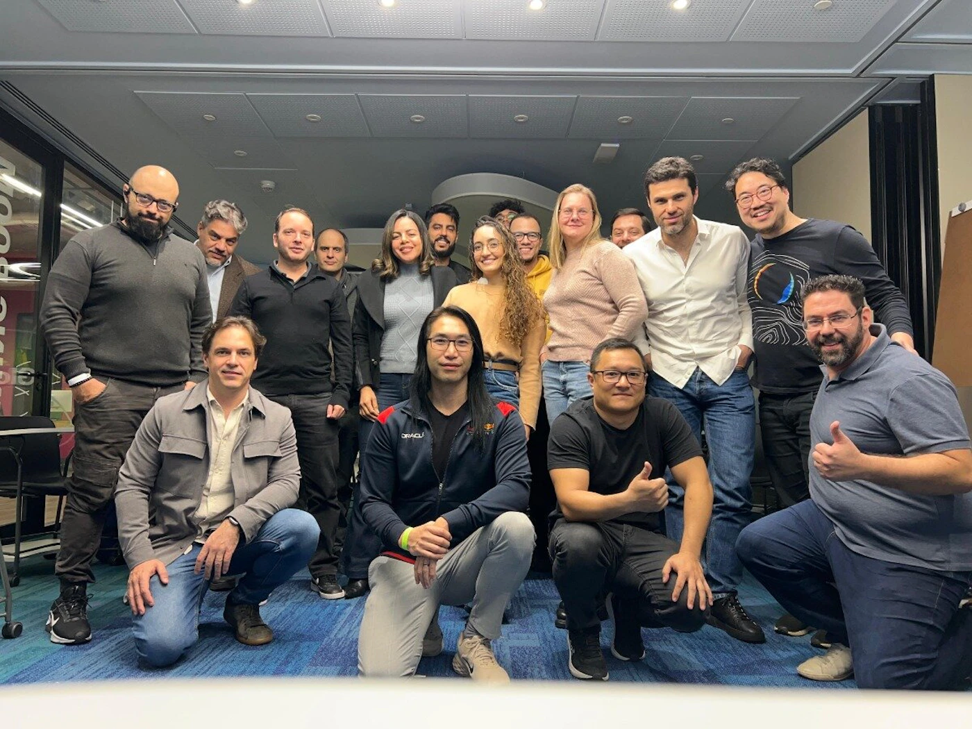 (Photo) A group of approximately eighteen diverse individuals, including men and women, are posing for a casual group photo indoors. They are smiling and looking towards the camera, with some standing and others kneeling in the front row. Text: ORACLE, Food group of people | indoor, office or conference space | diverse group, some kneeling, casual attire, blue patterned carpet | group portrait, candid | Colors: #4F4F4F, #8C8C8C, #336699, #D4B89B, #FFFFFF Note: This image is a direct capture of real-world subjects (people) in a specific setting, characteristic of a photograph rather than a graphic design or illustration.