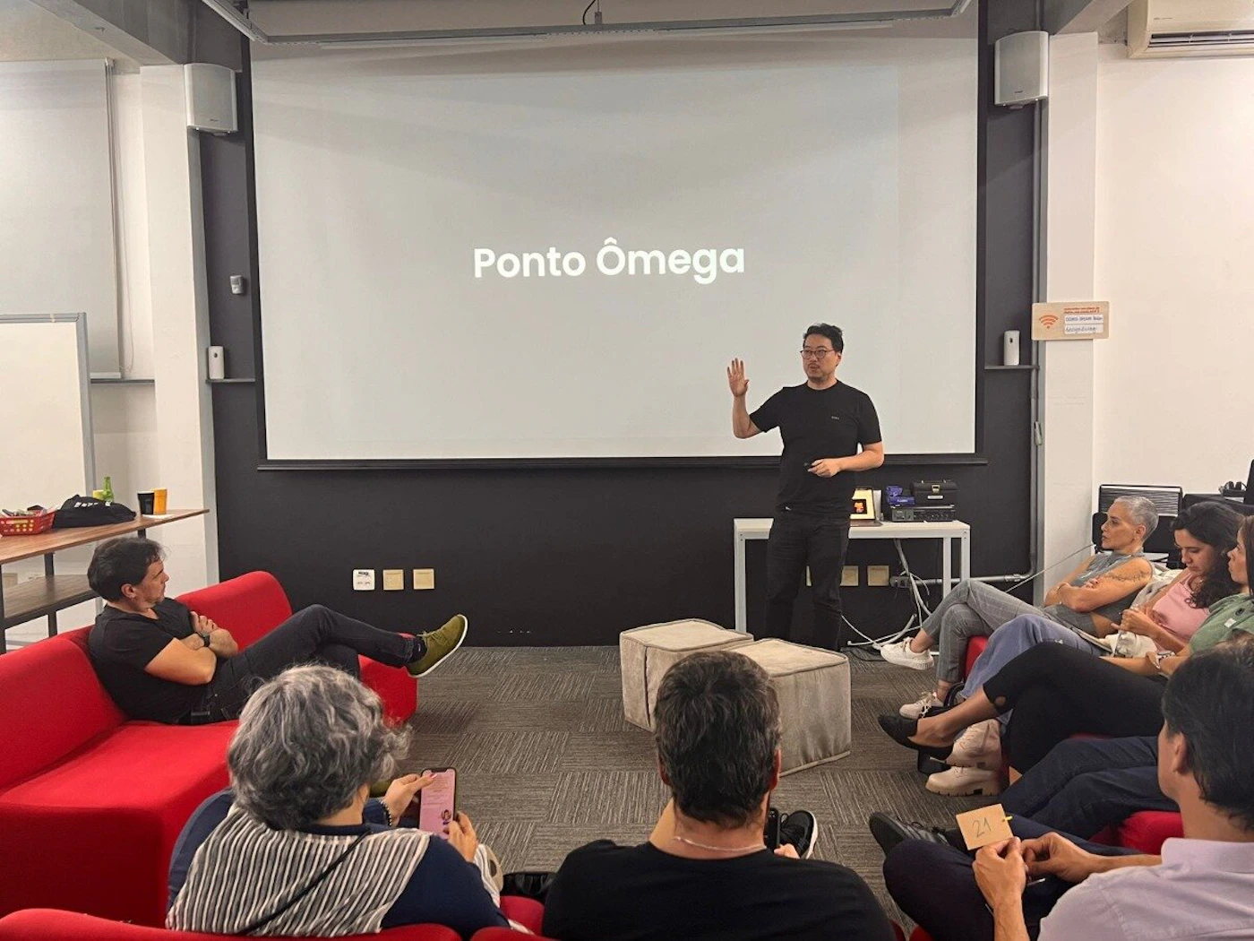(Photo) The image captures an indoor presentation or meeting where a man is standing in front of a projection screen displaying 'Ponto Ômega' and speaking to an audience seated on red sofas and floor poufs. The setting appears to be a modern office or co-working space. Text: Ponto Ômega Wi-Fi Grátis Rede: [unreadable] Senha: [unreadable] 23 people, presentation, meeting | indoor, modern office/meeting room | a presenter, an audience, a large projection screen, red sofas, Wi-Fi sign | candid, event photography | Colors: #E52E2E, #1A1A1A, #F0F0F0, #8C8C8C Note: This image is a direct capture of a real-world scene involving people, objects, and an environment, characteristic of a photograph rather than a designed graphic, illustration, or text-heavy document.