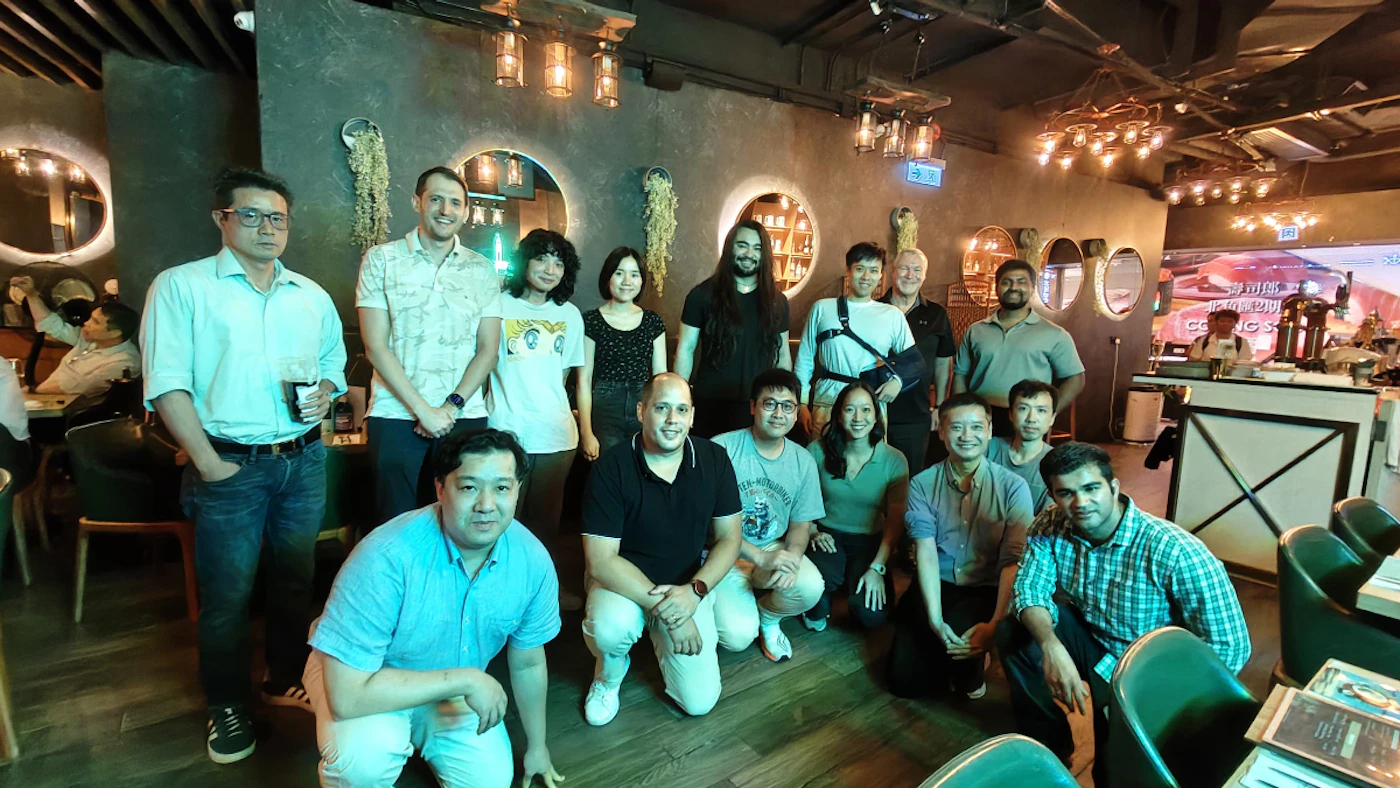 (Photo) A group of approximately fifteen people, appearing to be adults, are posing for a photo in a dimly lit indoor setting, likely a restaurant or bar. They are arranged with some standing and others crouching in the front, amidst decorative lighting, mirrors, and seating. Text: Coming S[unreadable], [unreadable Chinese text] A group of people | Indoor, restaurant/bar | Multiple people, dimly lit environment, decorative lighting, mirrors on walls, casual attire | Group portrait/candid | Colors: #363E3D, #F2D19F, #4A8B78, #E0E0E0 Note: This image is a photograph capturing a moment with real people in a real-world setting, rather than a graphic design, illustration, or text-based document.