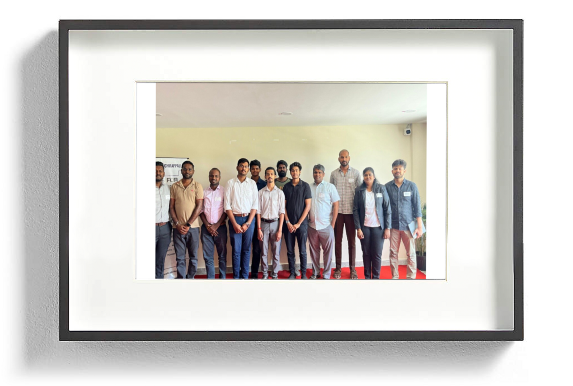 (Photo) A framed photograph depicts a group of approximately 12-13 individuals, predominantly men, standing in a line indoors, likely posing for a group picture. In the background on the left, a partially visible banner or board with text can be seen. Text: CORRUPTION CHAPTER FOR FLR group of people | indoor, likely a room or hall with a plain wall and red carpet | approximately 12-13 individuals, one woman among mostly men, standing in a line, partially visible text banner in the background | group portrait | Colors: #E0DCCA, #A01010, #303030, #F0F0F0 Note: The image displays a realistic depiction of a group of people, captured by a camera, which is characteristic of a photograph. It is not an illustration, logo, banner (though there's a banner *within* the photo), or handwriting.