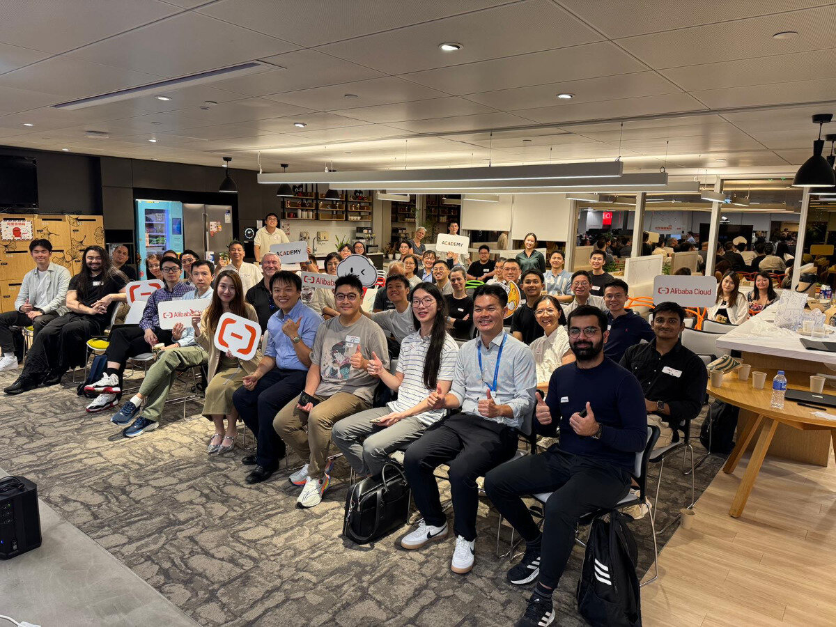 A group of approximately 30-40 people are seated indoors in what appears to be an office or event space, posing for a photograph. Many individuals are smiling, and some are holding white signs featuring the Alibaba Cloud logo and text.. Text: Alibaba Cloud ACADEMY