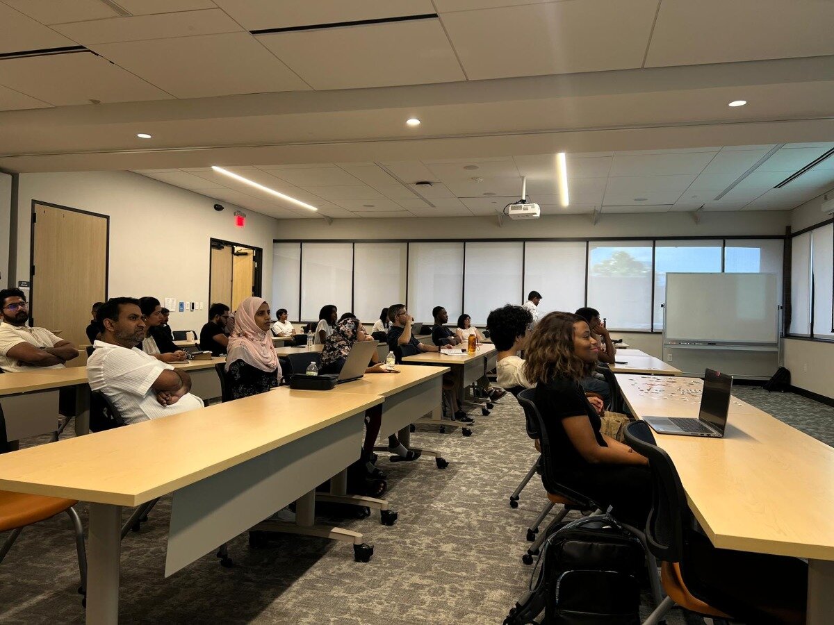 (Photo) A group of diverse individuals are seated at light-colored desks in a modern classroom or conference room, attentively facing towards the front where a projector screen and a whiteboard are visible. people attending a session or lecture | indoor, modern classroom or conference room | multiple individuals, some with laptops, a projector displaying a faint image, a whiteboard, and large windows with blinds | candid, wide-angle shot of a group | Colors: #DDC39B, #E0E0E0, #878A8D, #282828, #A7816A Note: The image is a realistic capture of a real-world scene, depicting people and an environment as they appear in life, which aligns with the definition of a 'photo'.