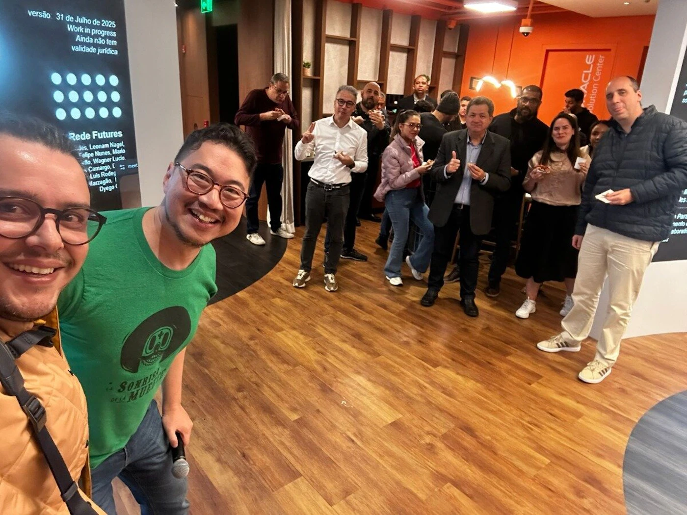 (Photo) The image captures a group of people, predominantly men, gathered indoors in what appears to be an office or event space. Two individuals in the foreground are smiling at the camera, while others in the background are standing and interacting, with some looking towards the camera. Text: versão 31 de Julho de 2025 Work in progress Ainda não tem validade jurídica Rede Futures Juss, Leonam Nagel Felipe Nunes, Mario [unreadable] Wagner Lucio Camargo, [unreadable] Luis Rodes Marzagão, Dyego ORACLE Solution Center Sonrisa de la Muerte people | indoor | group of people, some posing for a selfie, office/event space setting, large screen with text, corporate branding (Oracle) | candid, group photo | Colors: #8B4513, #FFA500, #4CAF50, #36454F, #F5F5DC Note: This image is a direct capture of a real-world scene featuring people and an environment, characteristic of a photograph. It is not an illustration, logo, banner, or handwriting sample.