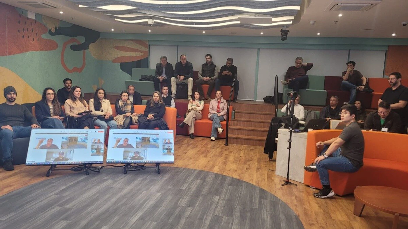 (Photo) A wide-angle photo captures a group of people attending a hybrid meeting or presentation in a modern, multi-level room, with some participants viewing video calls on two large screens in the foreground. The room features tiered seating, colorful couches, and a distinctive abstract wall mural. group of people, meeting, interior space | indoor, modern office or collaborative space | multiple people, tiered seating, large screens displaying video calls, abstract wall mural, unique ceiling design | candid, wide-angle shot | Colors: #E8743B, #80C8B8, #8A6D52, #5A606A Note: The image is a realistic capture of a physical environment and people, showing a real-world scene rather than a graphic design, illustration, or text-based content.