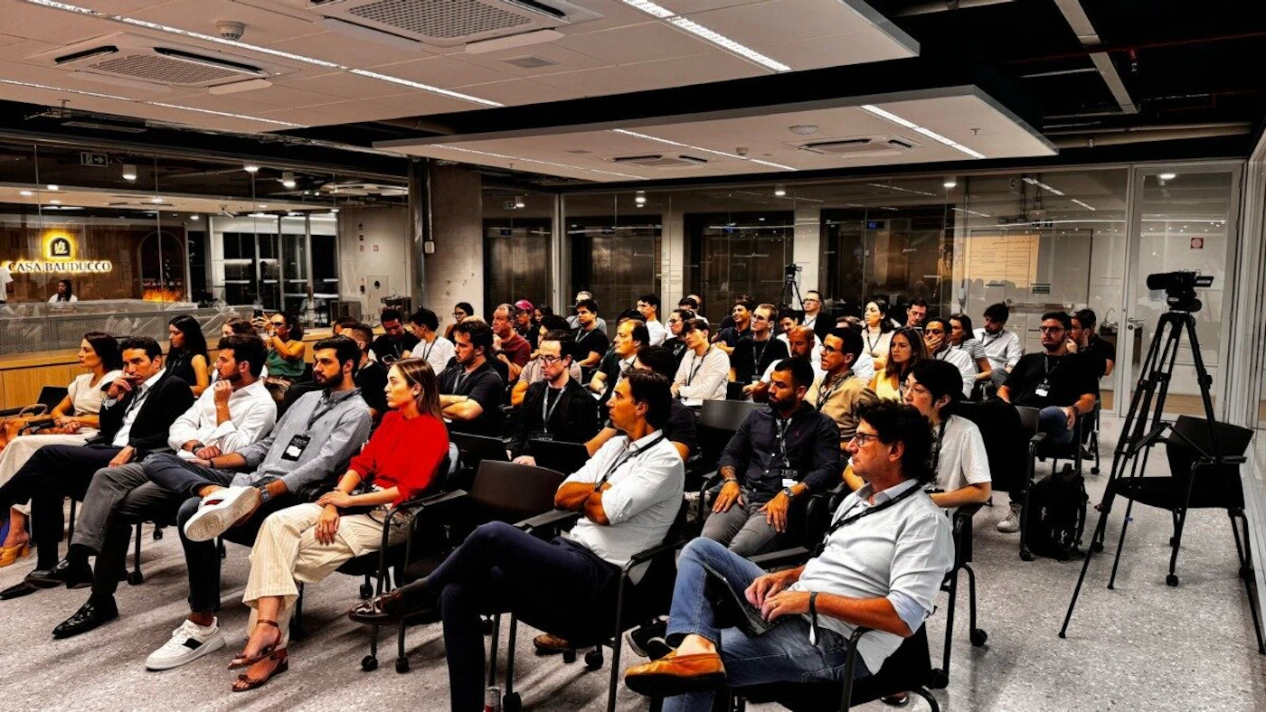 (Photo) A wide-angle photograph captures a large group of people, mostly adults, seated in an indoor conference or event space, attentively facing forward. A camera on a tripod is visible on the right side of the frame. Text: CASA BAUDUCCO People attending an event or presentation | Indoor, conference room or auditorium | Multiple people seated in rows, a professional camera on a tripod, a sign for 'CASA BAUDUCCO' in the background, modern interior design with glass partitions and exposed ceilings. | Candid, event photography | Colors: #2C2C2C, #E0E0E0, #A07040, #E04040, #507090 Note: This image is a direct capture of a real-world scene, featuring multiple individuals and an indoor environment, which aligns with the definition of a photograph.
