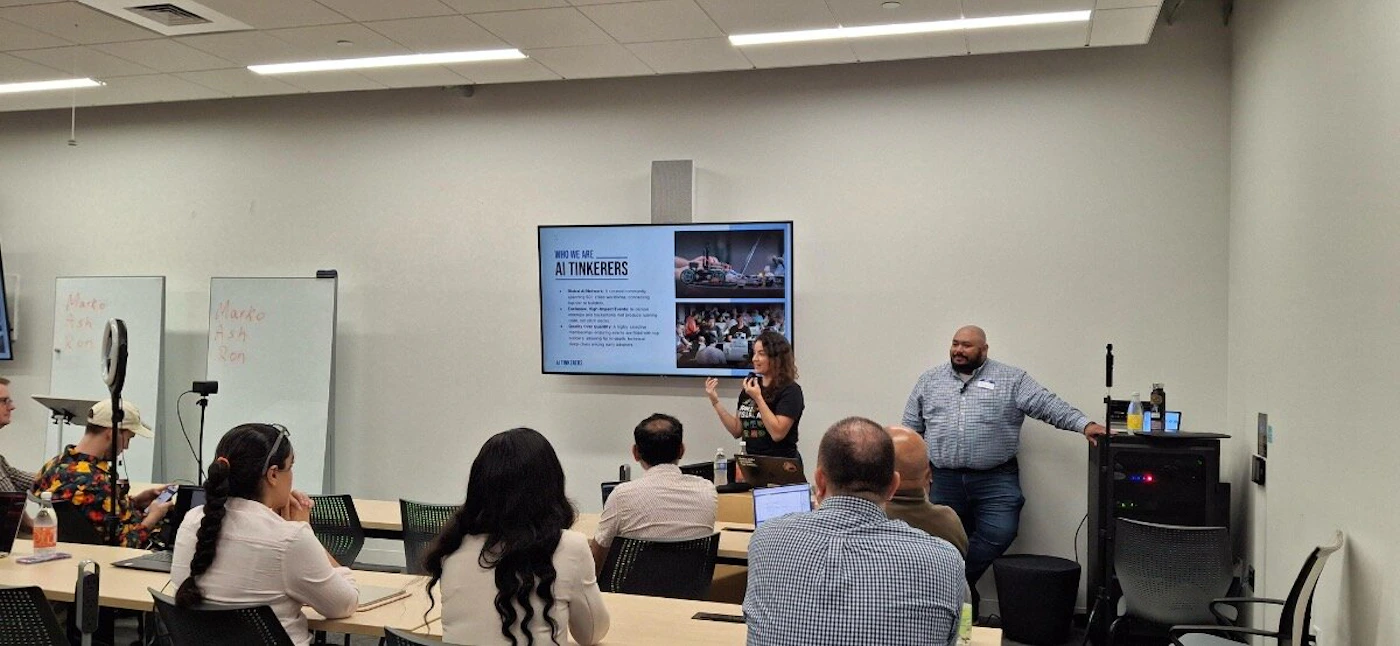 (Photo) A photograph captures an indoor presentation or workshop where a woman stands near a large screen displaying a slide titled 'WHO WE ARE AI TINKERERS' while speaking to an audience seated at tables. Text: WHO WE ARE AI TINKERERS [Multiple lines of descriptive text] [Logo text] Maite Tom Ron Marko Ash Ron Group of people attending a presentation/workshop | Indoor (conference room/classroom) | Presentation screen, whiteboards with handwritten names, multiple attendees, presenter speaking, ring light setup. | Candid, wide shot | Colors: #FFFFFF, #D6C1AE, #000000 Note: The image is a candid photograph documenting a real-world event (a business or educational presentation) taking place in a conference room setting.