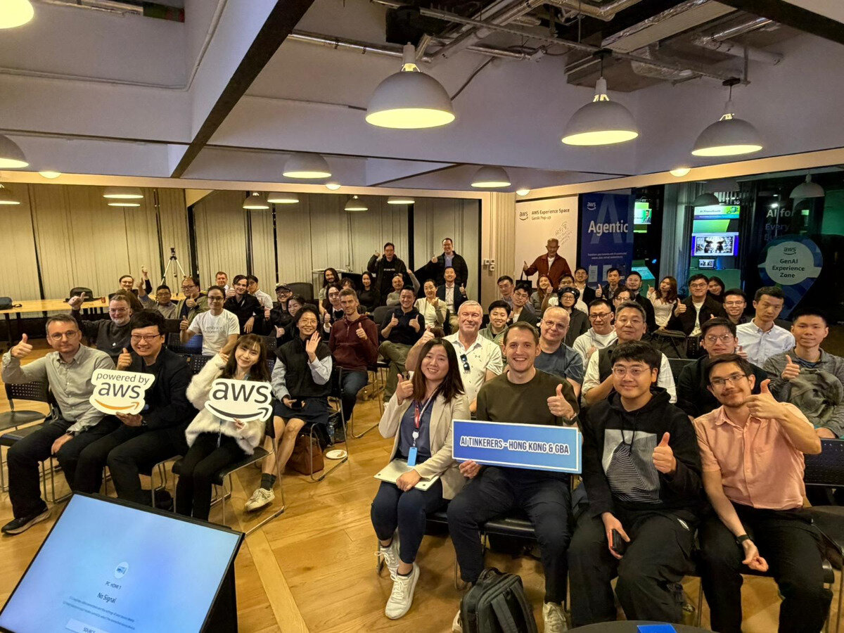 A photograph capturing a large group of people attending an indoor event, likely related to AWS and AI, all smiling and giving thumbs-up gestures.. Text: powered by aws aws AI TINKERERS - HONG KONG & GBA aws self-experience zone Agentic AI for Every AWS GenAI Experience Zone PC-HDMI 1 No Signal