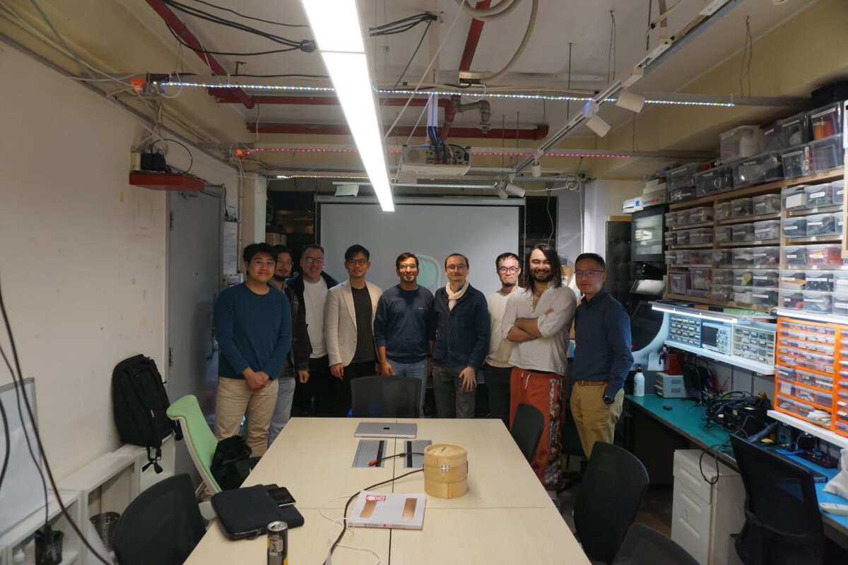 A group portrait featuring nine men standing behind a conference table in what appears to be a brightly lit, cluttered workshop or lab environment.