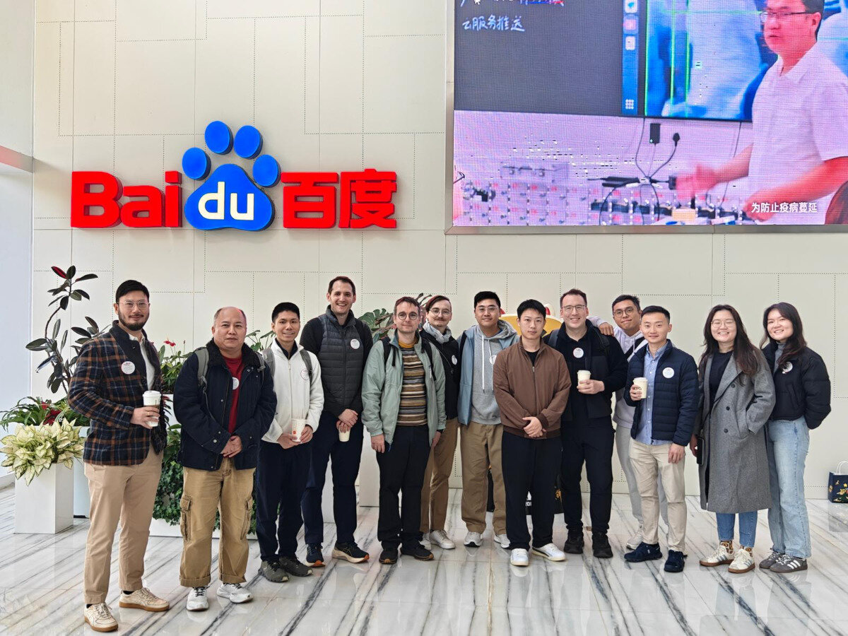 A group photo of approximately thirteen people, appearing to be colleagues or visitors, standing indoors in front of a large wall featuring the prominent red and blue Baidu logo and Chinese characters.. Text: Baidu 百度 工服务推送 为防止疫病蔓延