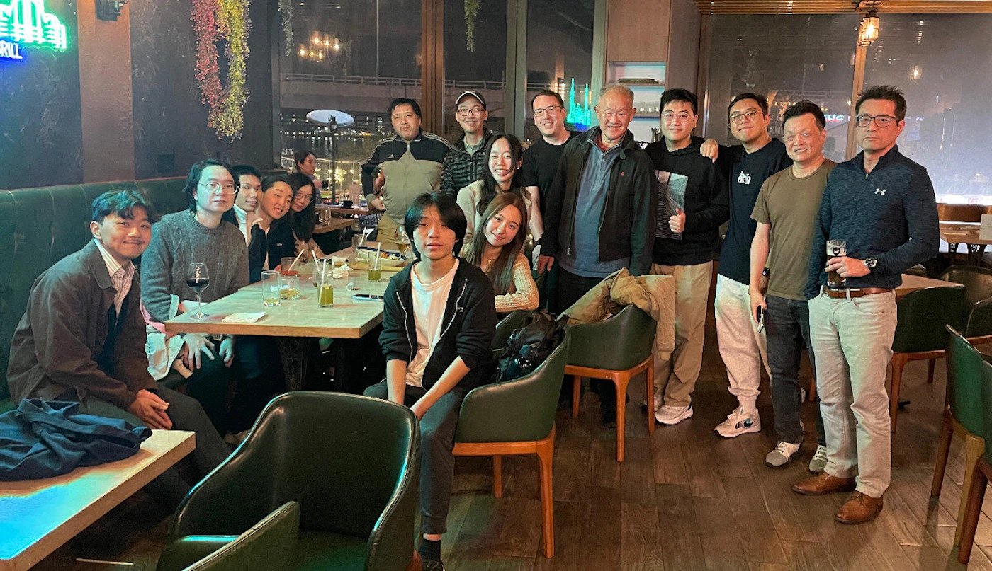 (Photo) A photograph capturing a large group of approximately 14 people posing for a group photo inside a dimly lit restaurant or bar during the evening. Text: [unreadable] GRILL Group of people (adults) | Indoor restaurant/bar, evening/night | Large group gathering, dark ambiance, green leather seating, neon sign visible in the background, large windows overlooking an urban area. | Candid/group portrait | Colors: #384f42, #1c1c1c, #a08269 Note: The image is a realistic photograph documenting a social gathering of a large group of people in an indoor dining setting.