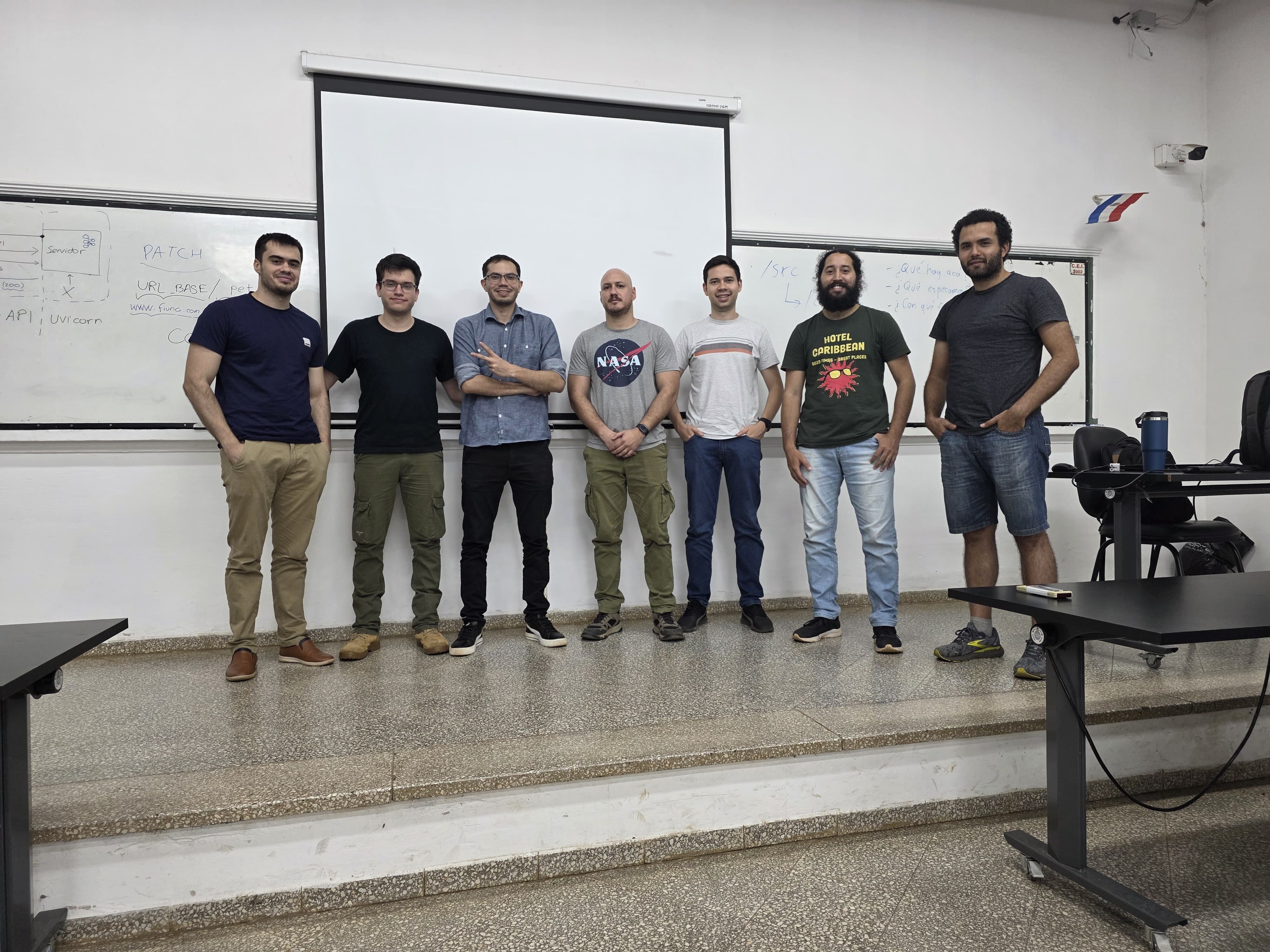 A group of seven men posing for a photo in front of a projector screen and a whiteboard in a classroom or office setting.. Text: PATCH URL_BASE / API API UNICORN NASA HOTEL CORIBBEAN BEST PRICE BEST SERVICE GREAT PLACES /src L...