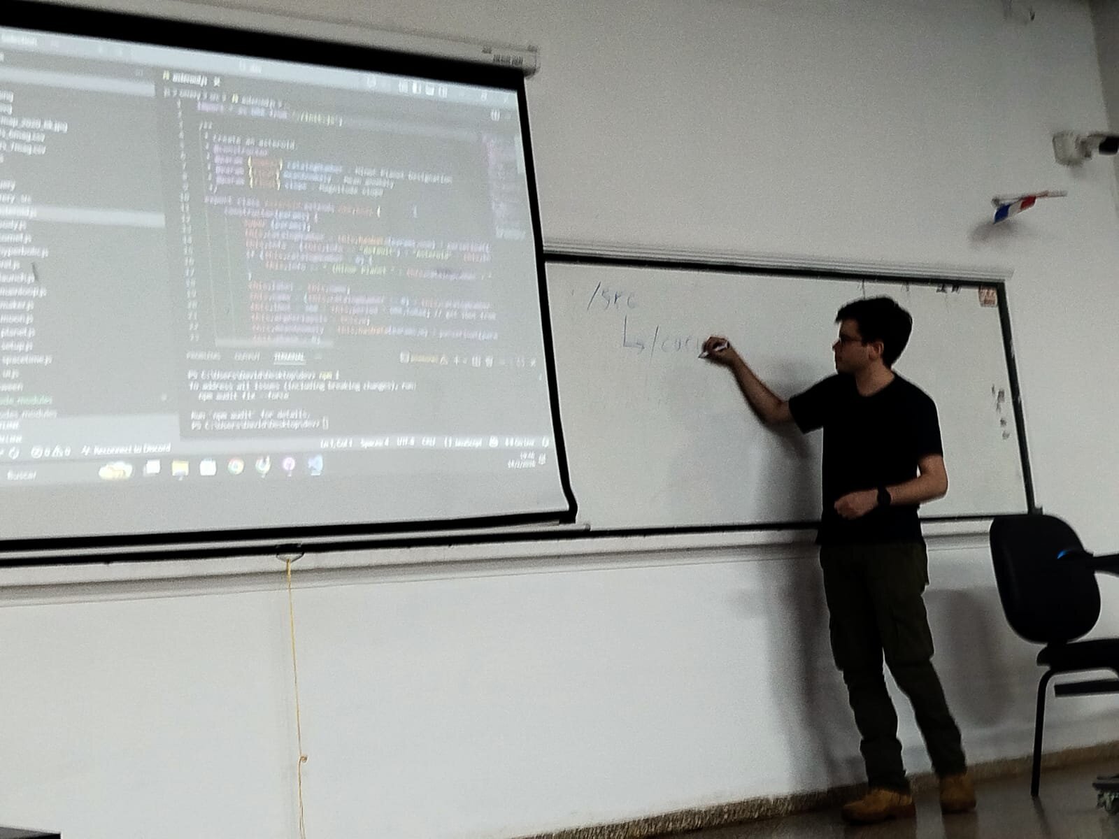 (Photo) A man stands in a classroom or lecture hall, writing on a whiteboard while a projector displays JavaScript code on a screen next to him. Text: import * as THREE from 'three'; /**  * Create an asteroid.  * @constructor  * @param {Object} catalogNumber - Minor Planet Designation  * @param {Object} discovery - Year, discovery  * @param {Object} slope - Magnitude slope  */ export class Asteroid extends THREE.Mesh {   constructor(params) {     super(params);     // ...   } } PS C:\Users\Dani\Desktop\dev> npm i to address all issues (including breaking changes), run:   npm audit fix --force Run 'npm audit' for details. PS C:\Users\Dani\Desktop\dev> /src   L /core person teaching or presenting | indoor classroom | projector screen with code, whiteboard with diagrams | candid | Colors: #FFFFFF, #000000, #333333 Note: The image is a photograph capturing a real-life scene of a person in an indoor environment.